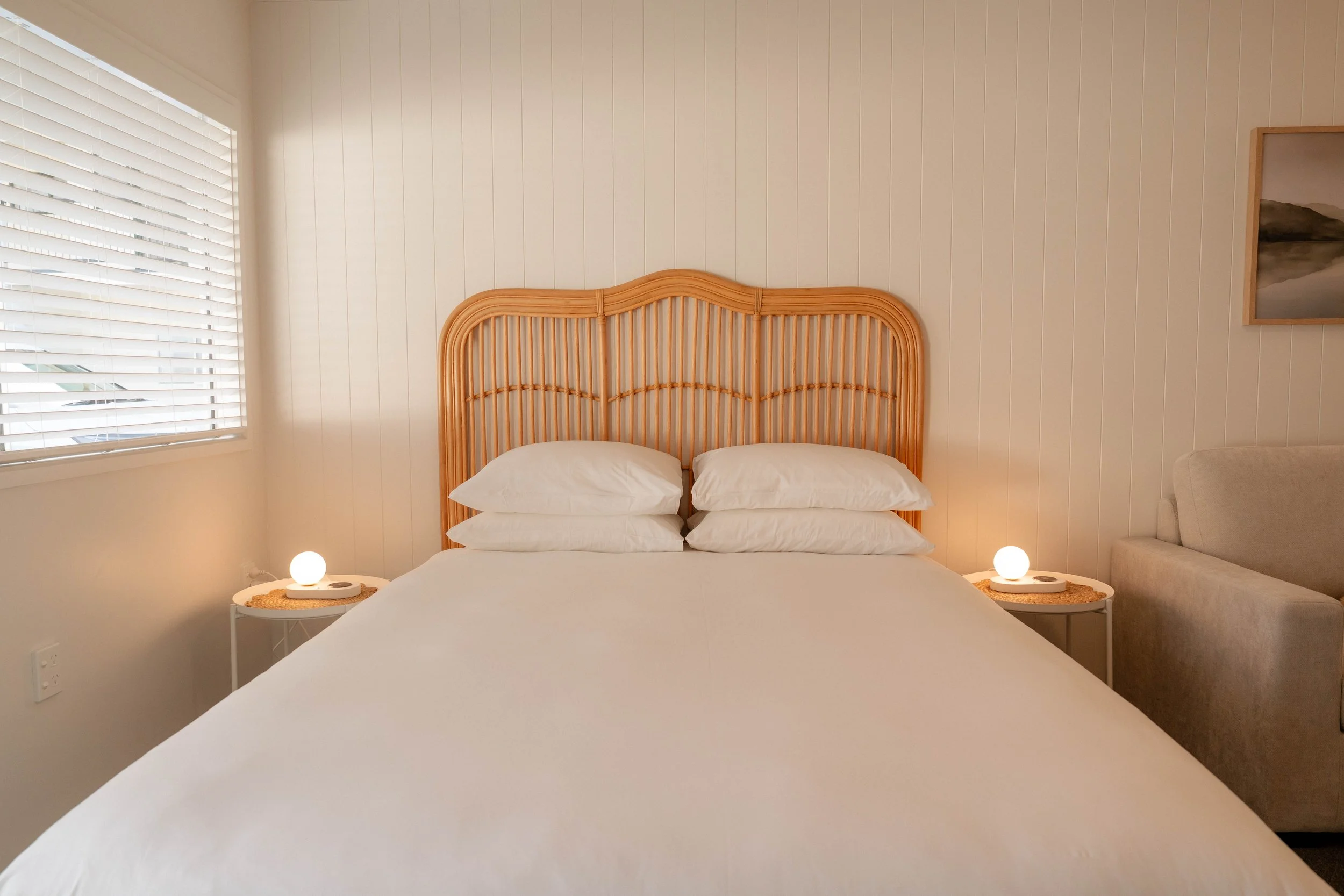 A bedroom with a bed featuring multiple white pillows, a wooden headboard, two round bedside lamps, a window with white blinds on the left, and a framed landscape painting on the wall to the right.