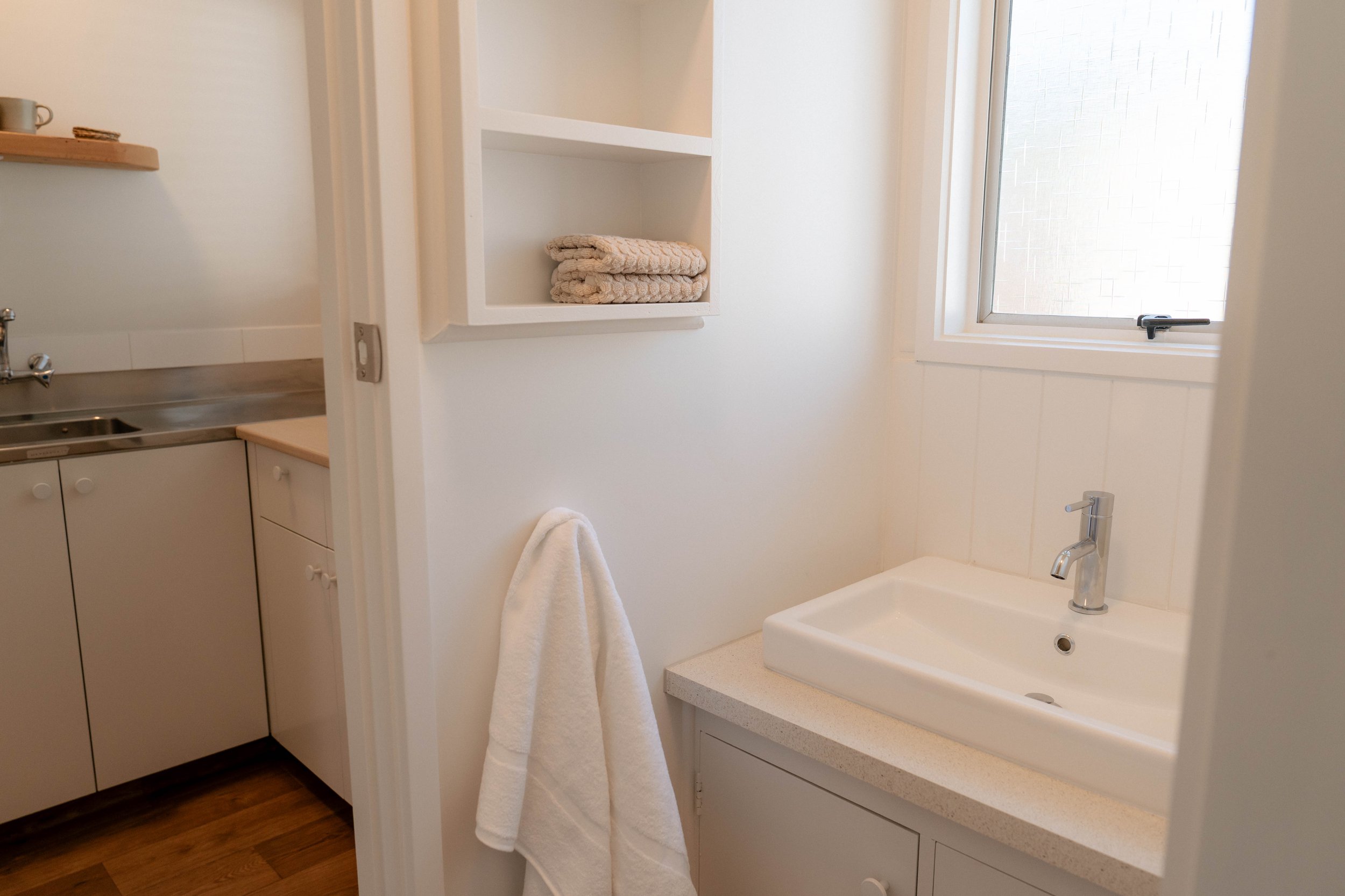 A bathroom corner with a white sink and faucet, a window with frosted glass, a towel hanging on a hook, and a built-in shelf with folded towels.