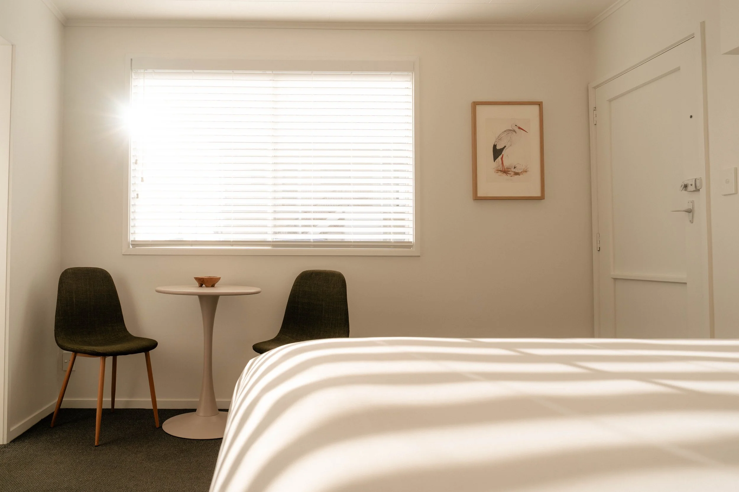 A hotel room with a window, two chairs, a small round table, a framed picture of a heron on the wall, and part of a bed.