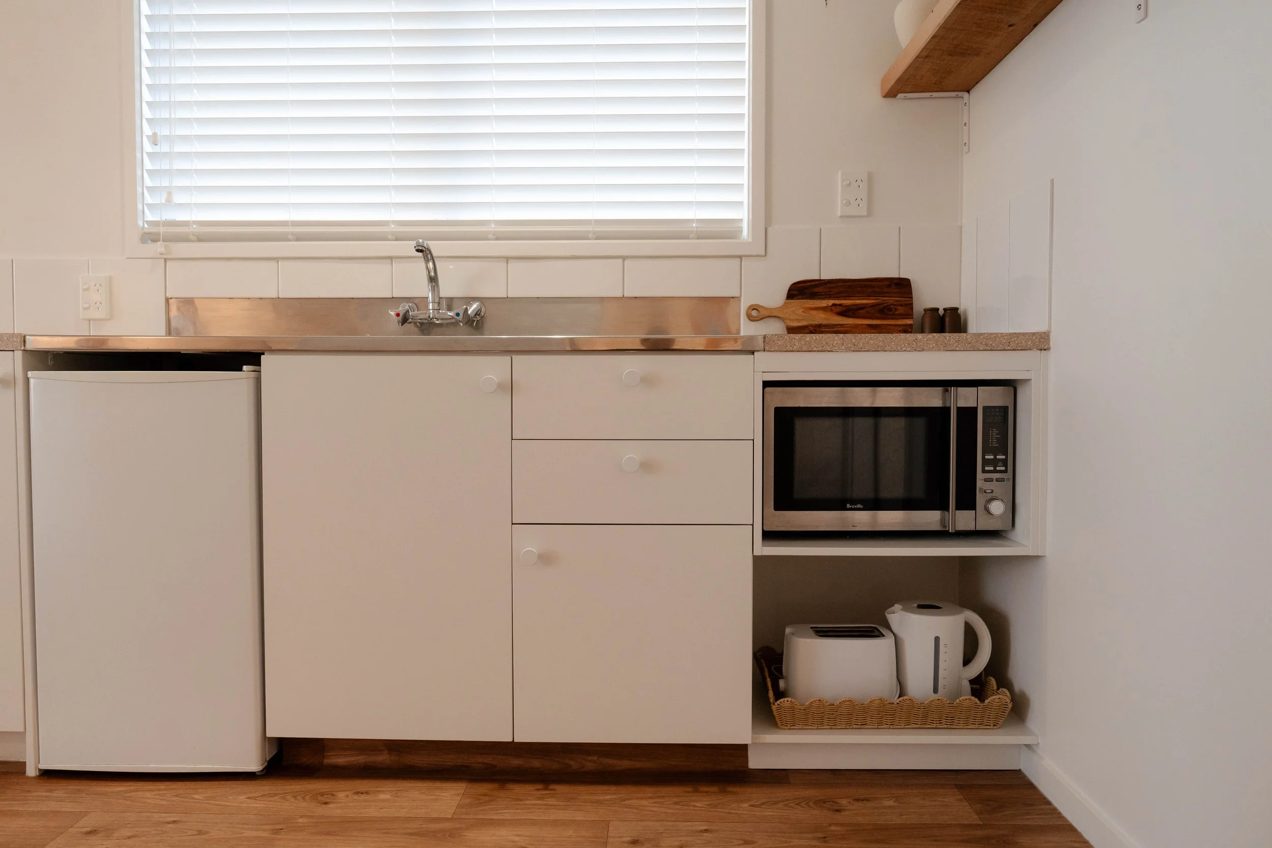 A kitchen counter with a sink underneath a window with closed white blinds. There are white cabinets below the counter, a microwave oven, a cutting board, and small containers on the countertop. A wicker basket and a water boiler are on the lower she