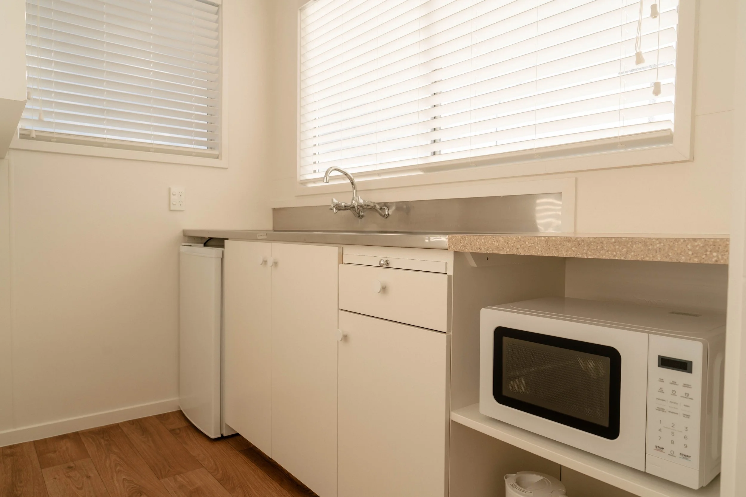 A small kitchen area with white cabinets, a countertop, a microwave oven, and a stove. Two large windows with white blinds let in natural light. There is a small refrigerator and part of a microwave oven. The floor is wood.