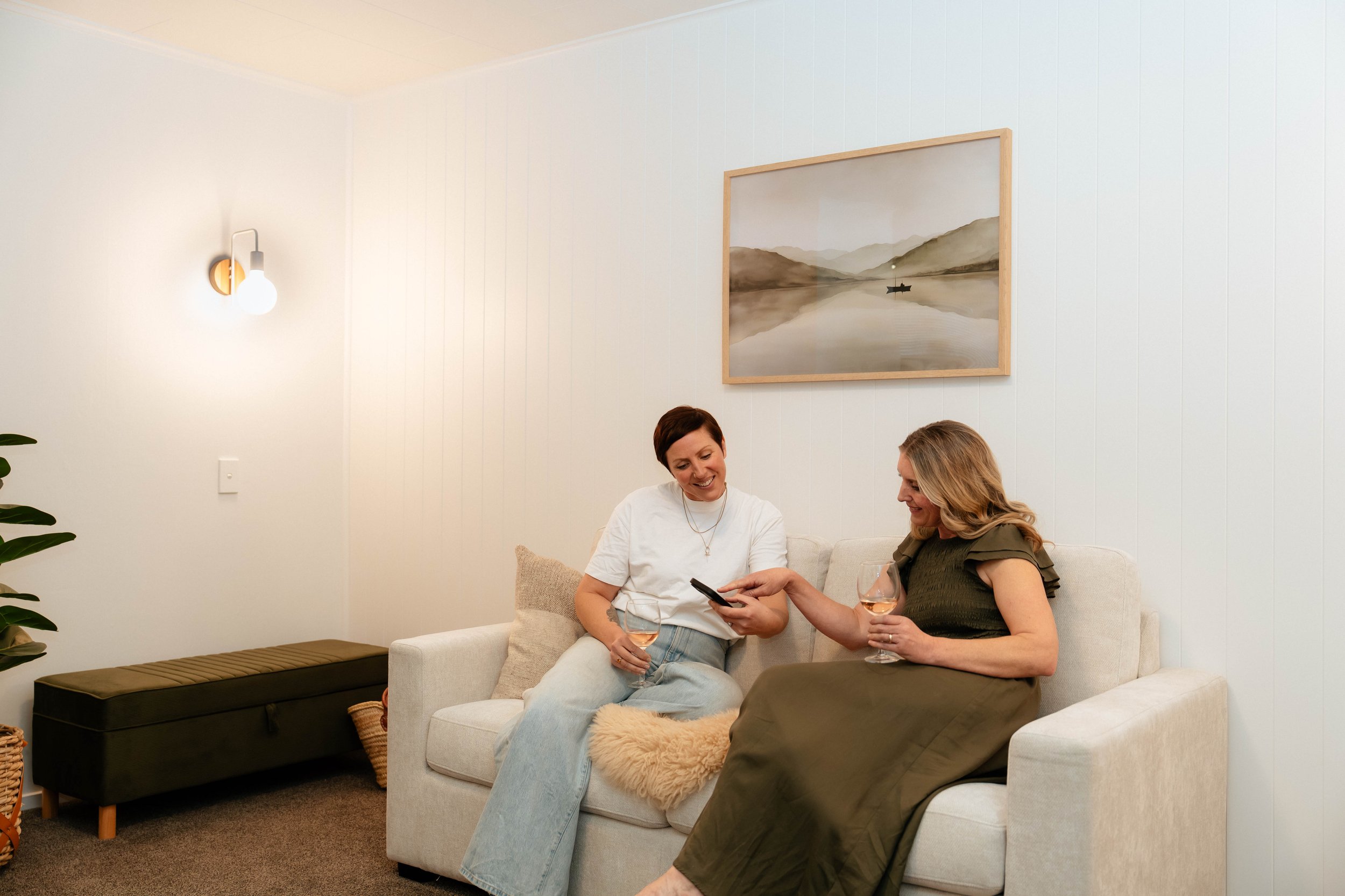 Two women sitting on a cream-colored sofa in a living room, smiling and looking at a smartphone together, each holding a glass of rosé wine. The room features minimalist decor with a landscape painting and a modern wall sconce on a white paneled wall.