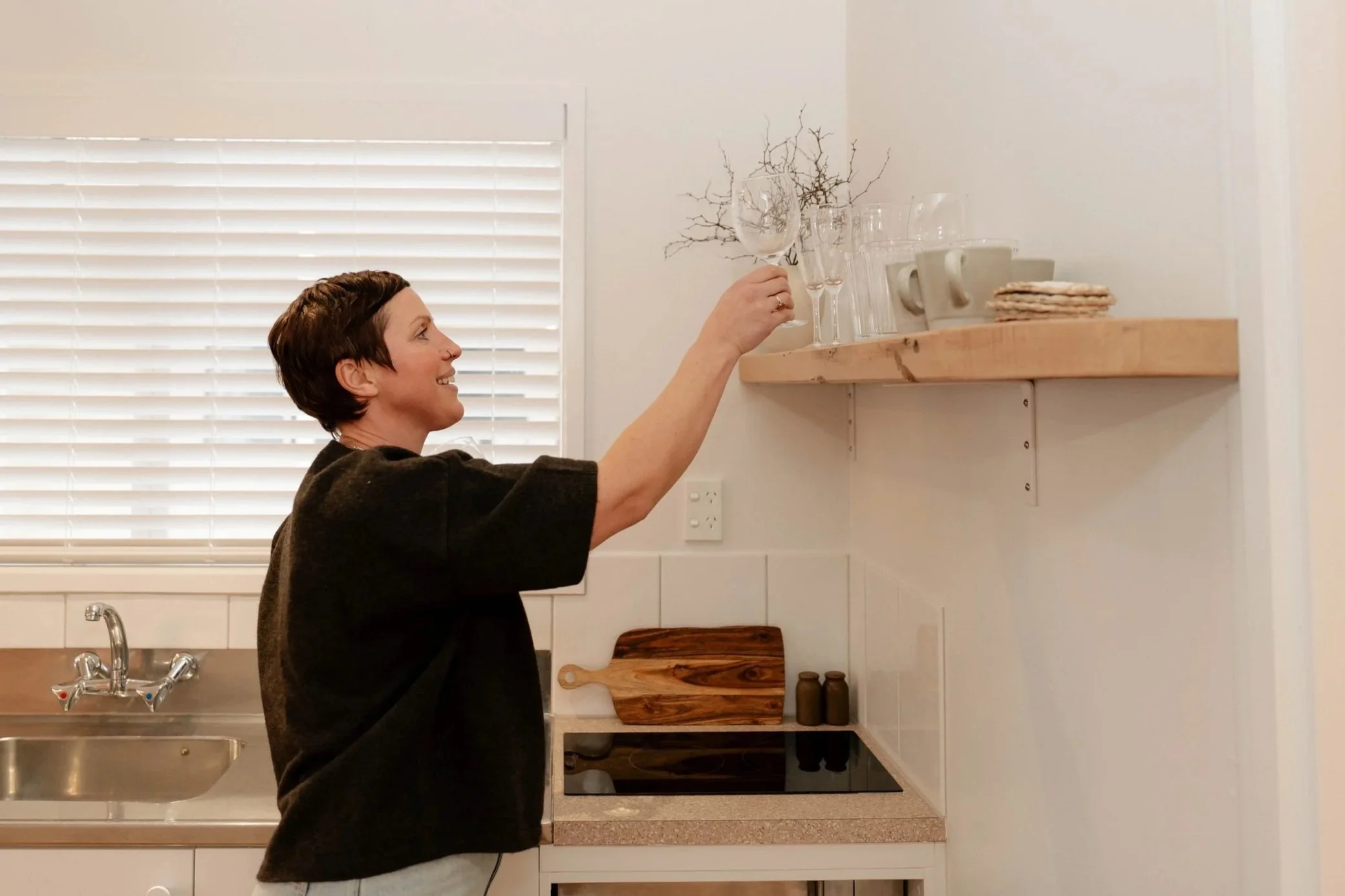 A woman with short dark hair holding a wine glass in a kitchen with white walls, wooden shelves, and blinds covering a window.