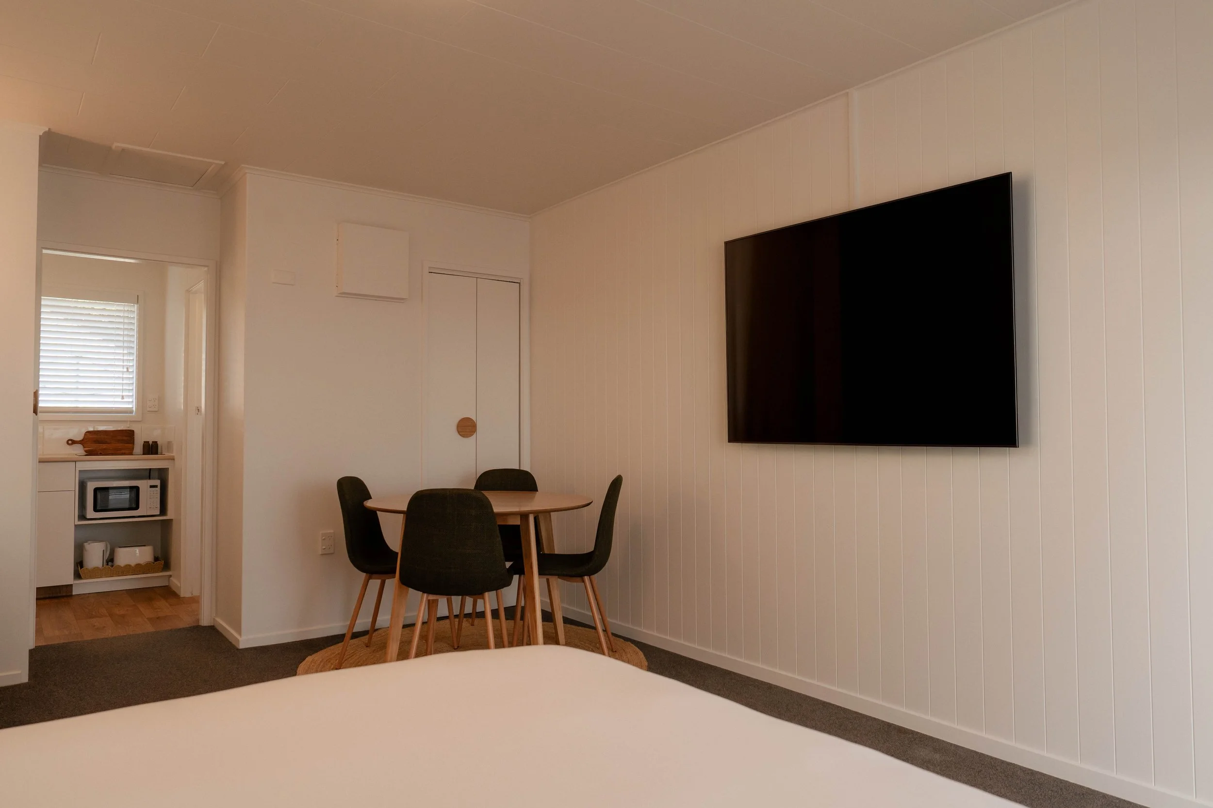 Interior of a small dining area with a round wooden table, four black chairs, a wall-mounted flat-screen TV, and a doorway leading to a kitchen with a microwave and window with blinds.