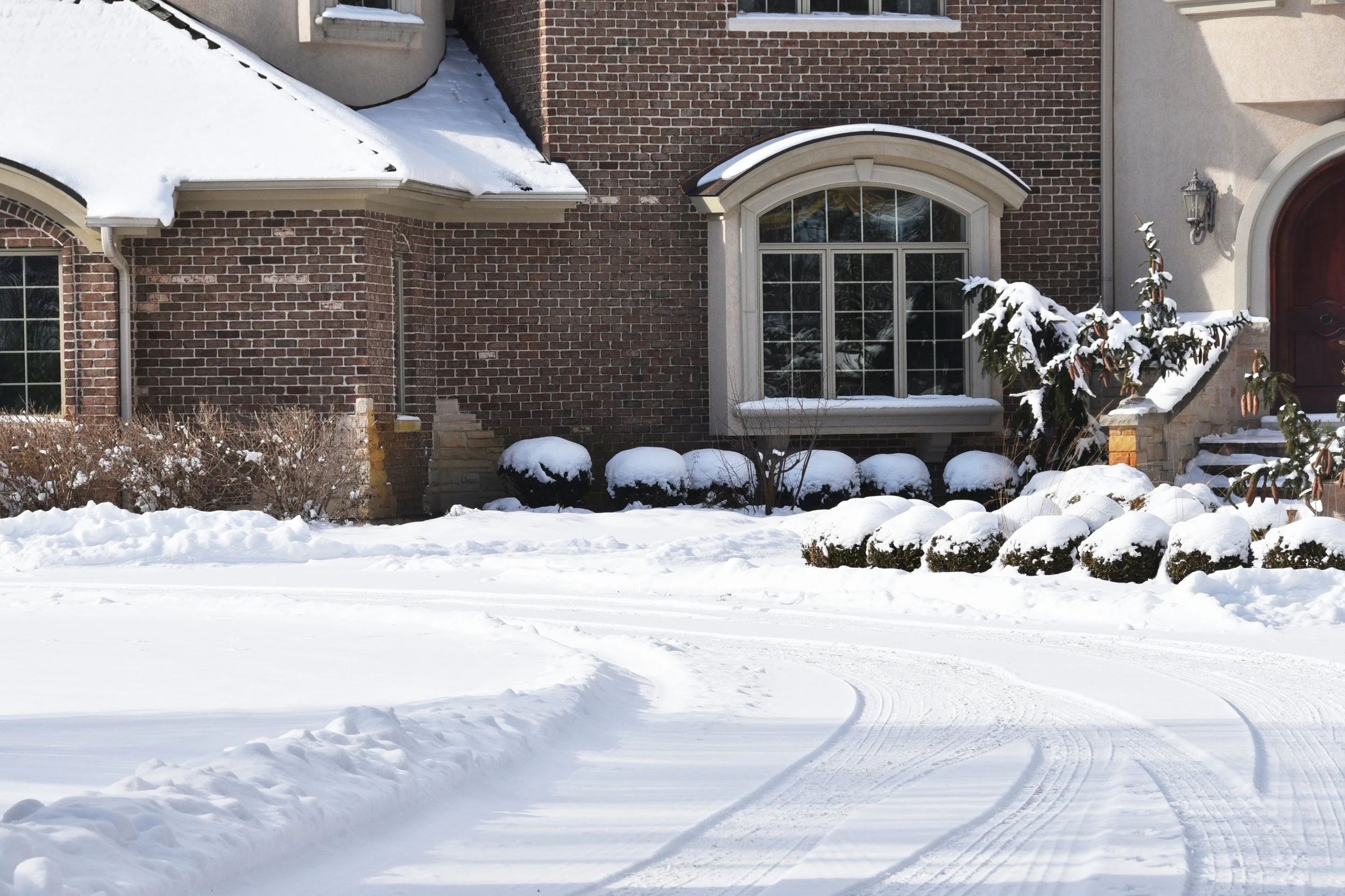 Front of a brick house with snow-covered roof, bushes, and driveway in winter.