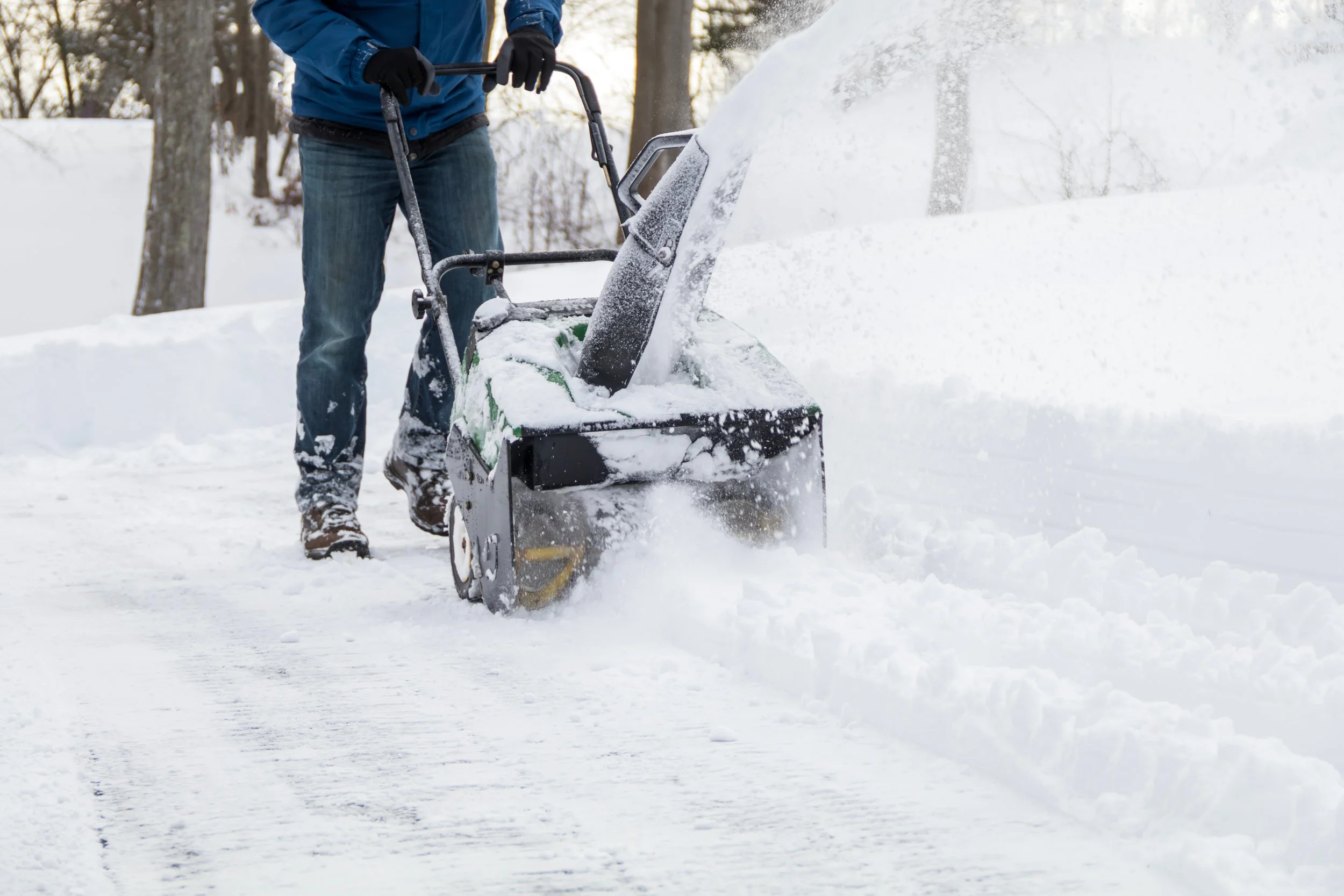 Person using a snow blower to clear snow from a driveway in a wintery wooded area.
