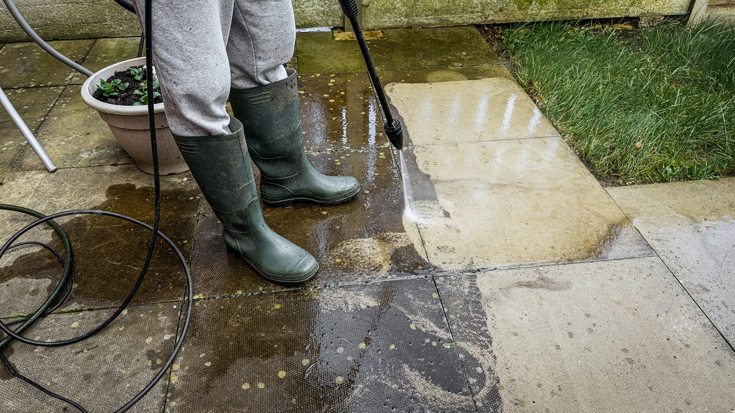 Person wearing gray sweats and green rubber boots pressure-washing a tiled patio, with a large pot of plants nearby and green grass at the edge of the patio.