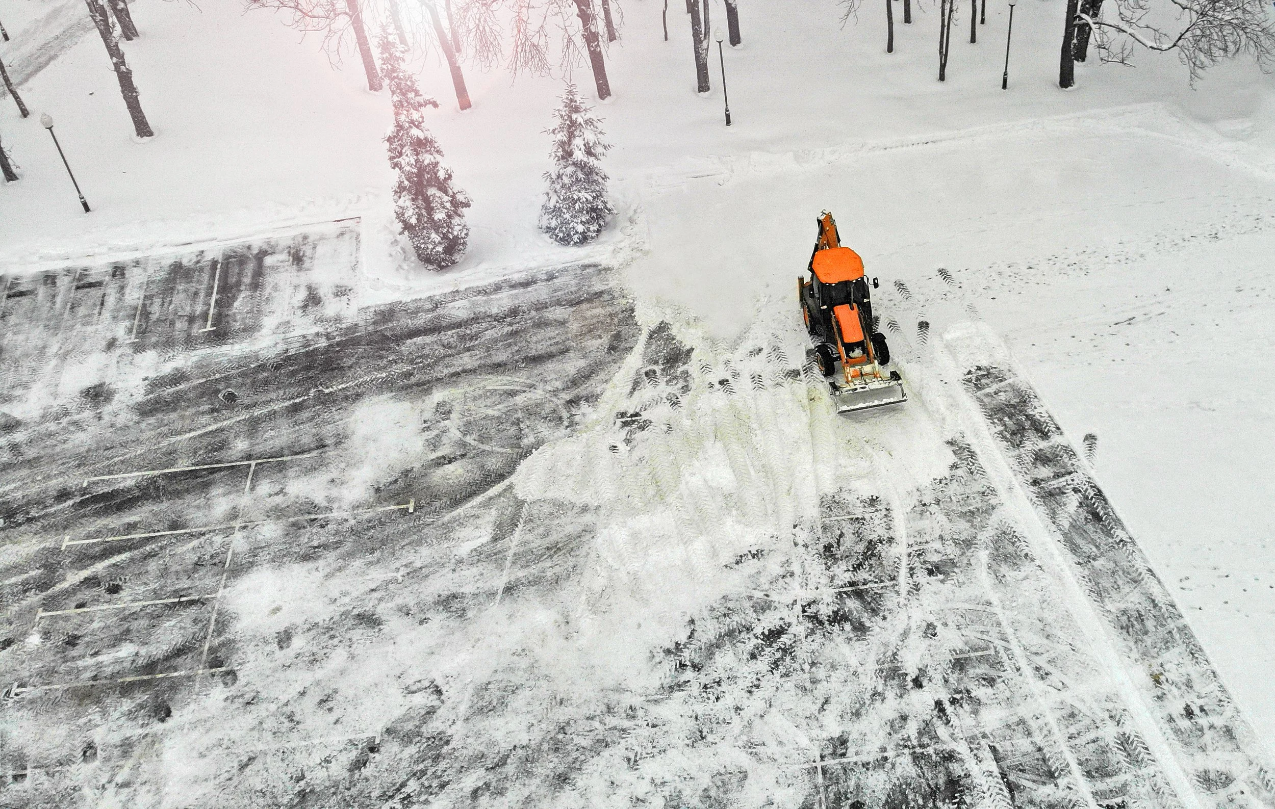 Aerial view of a snow-covered parking lot being cleared by an orange snow plow, with tire tracks and footprints visible in the snow.