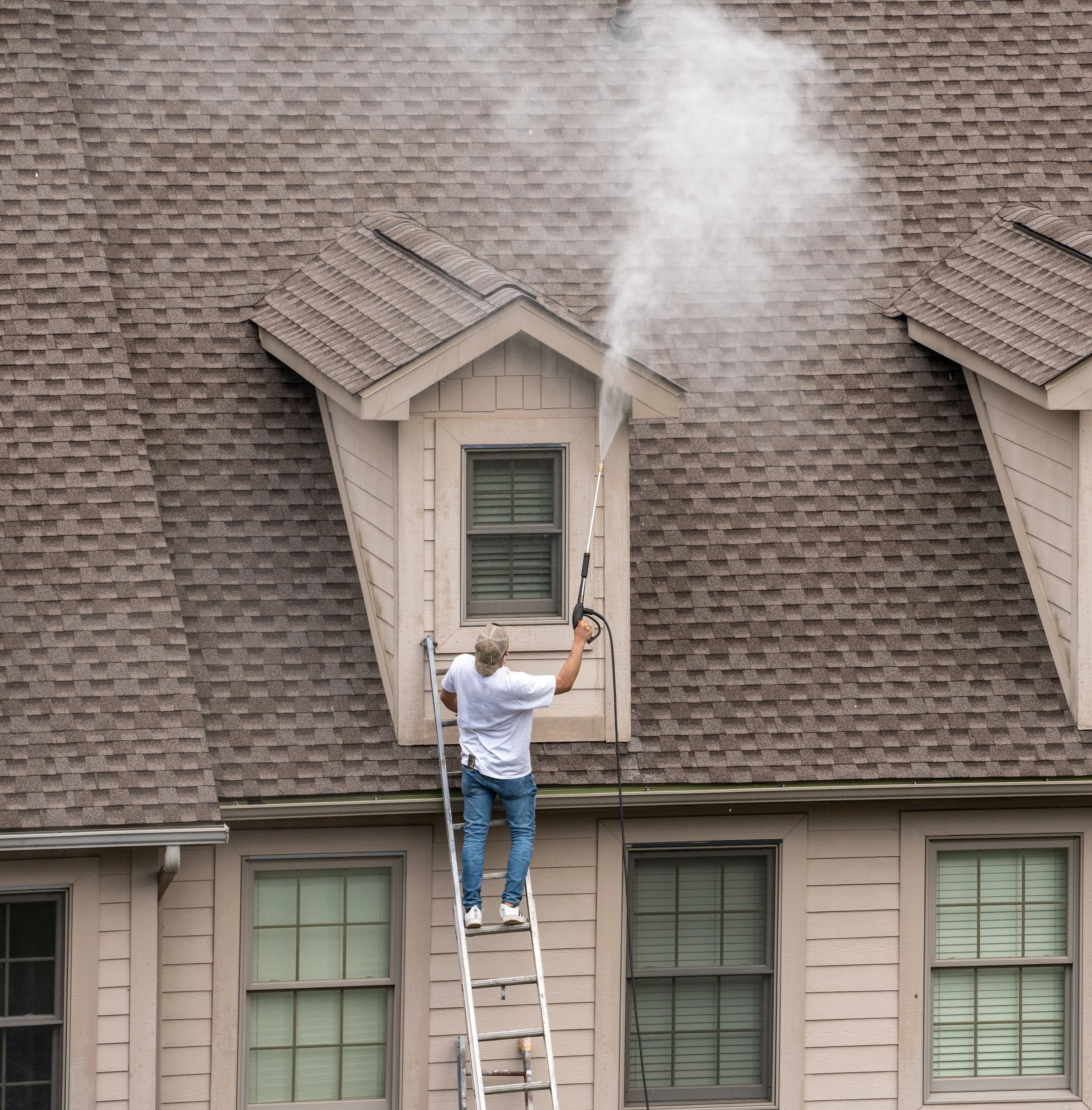 A man standing on a ladder cleaning a window on the second floor of a house with beige siding and a steep shingled roof, using a pressure washer.