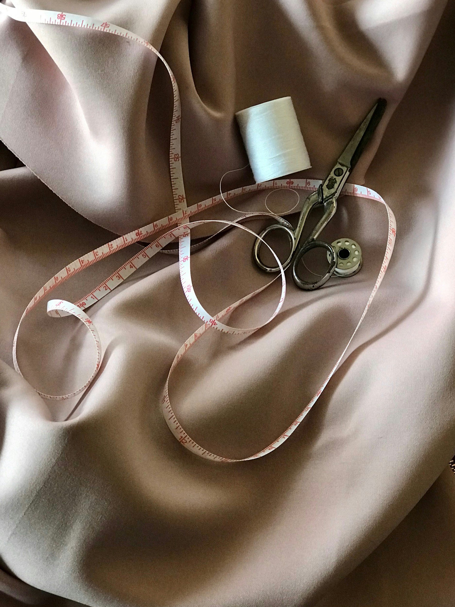Sewing materials on a shiny, light pink fabric background, including a white measuring tape, a spool of white thread, and a pair of vintage silver scissors.