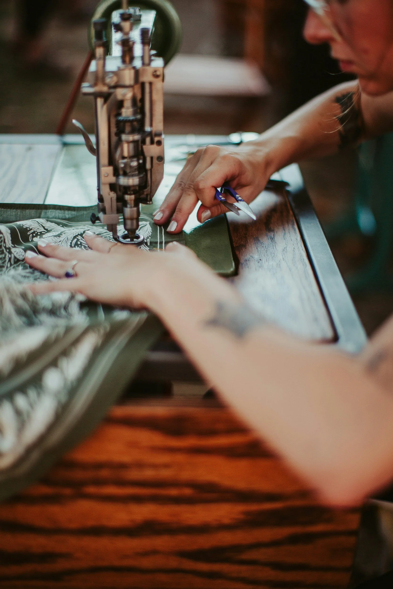 A person operating a vintage sewing machine, sewing a fabric with a floral pattern on a wooden table.