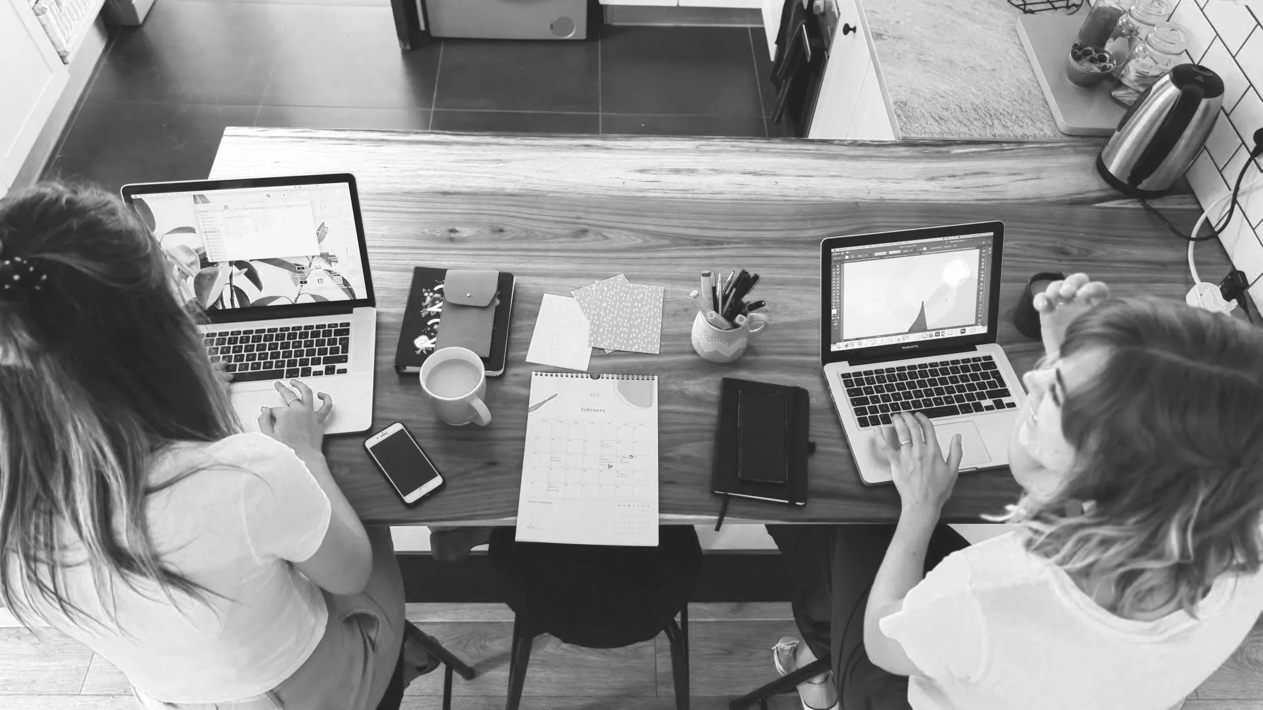 Two women working on laptops at a wooden table with notebooks, a calendar, a mug, a smartphone, pens, and a jar on the table in a kitchen.