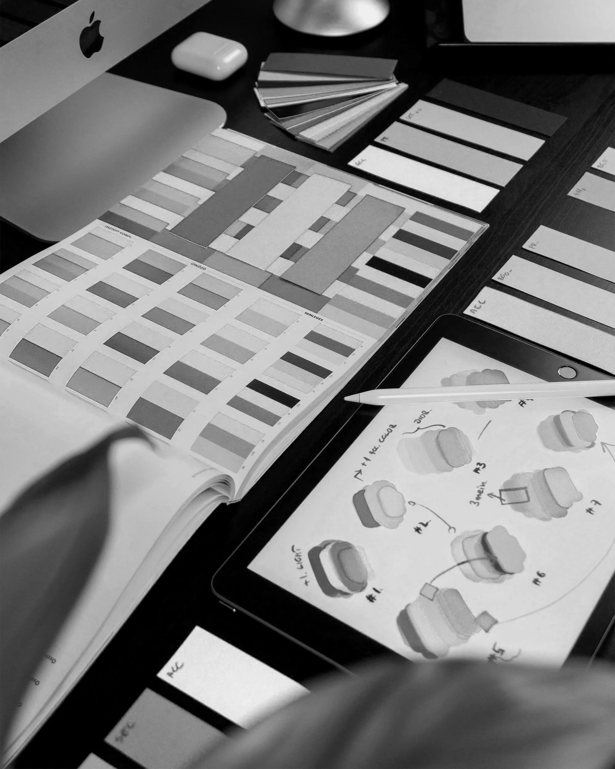 Black and white photograph of a workspace featuring color swatches, a palette with paint sample illustrations, a notebook, a pen, and a computer monitor.