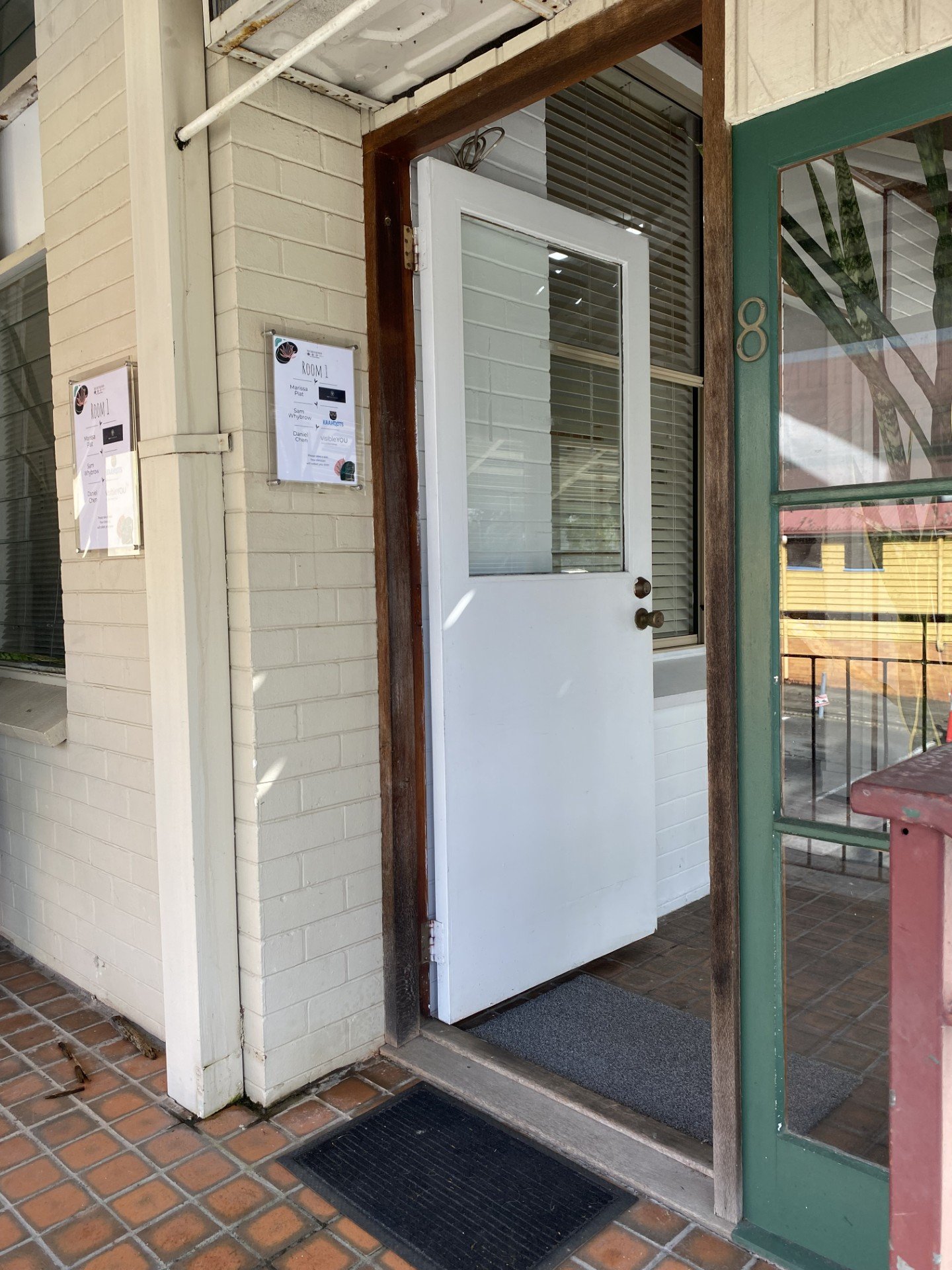 The door of the clinic. Open white door with window panes, leading into a building with green trim and brick floor, numbered 8, with a black doormat outside.