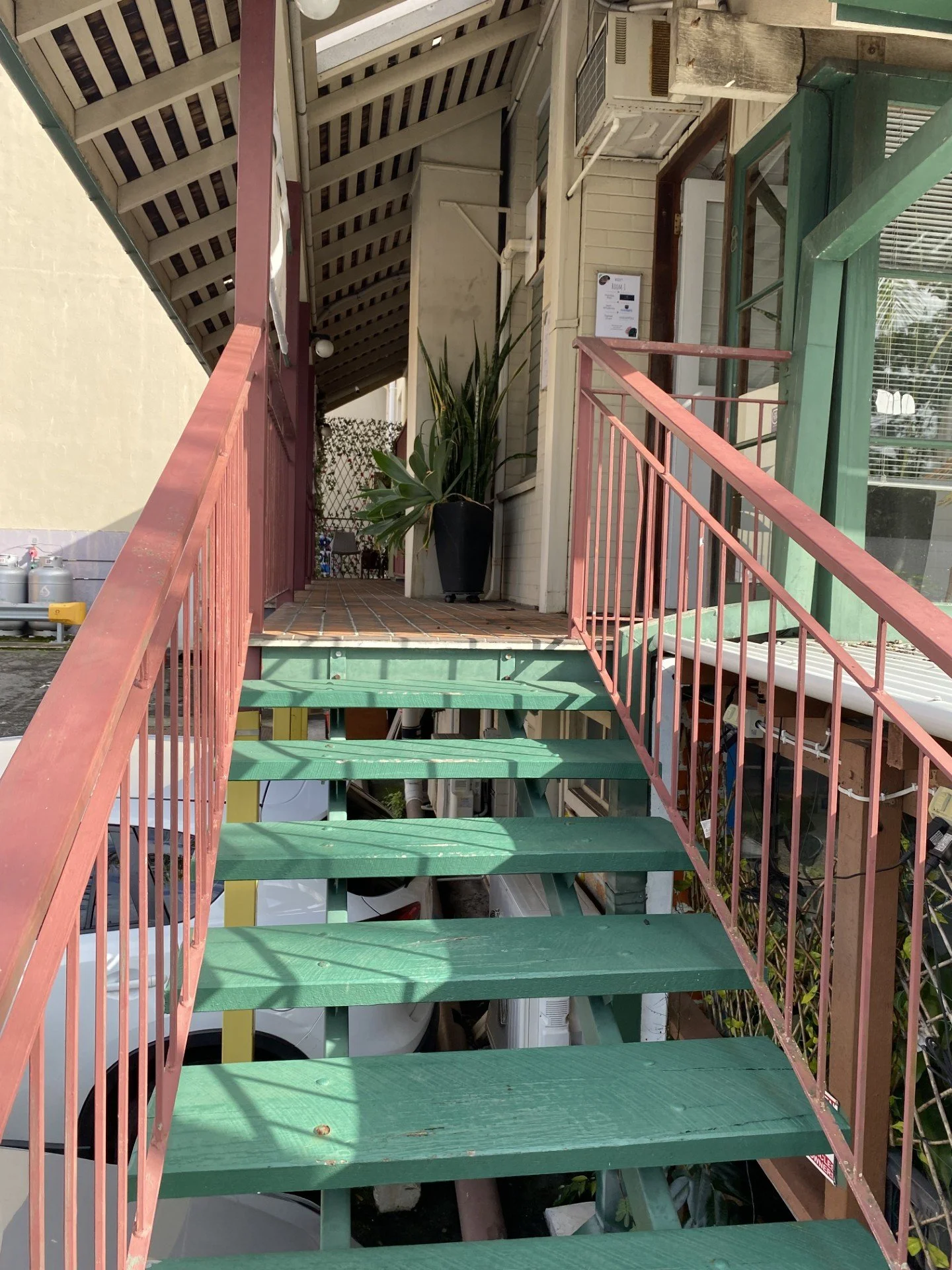 The stairs to the clinic. View of a multi-level outdoor staircase with green steps and pink railings leading up to a balcony with potted plants, windows, and an air conditioning unit on a building's exterior wall.