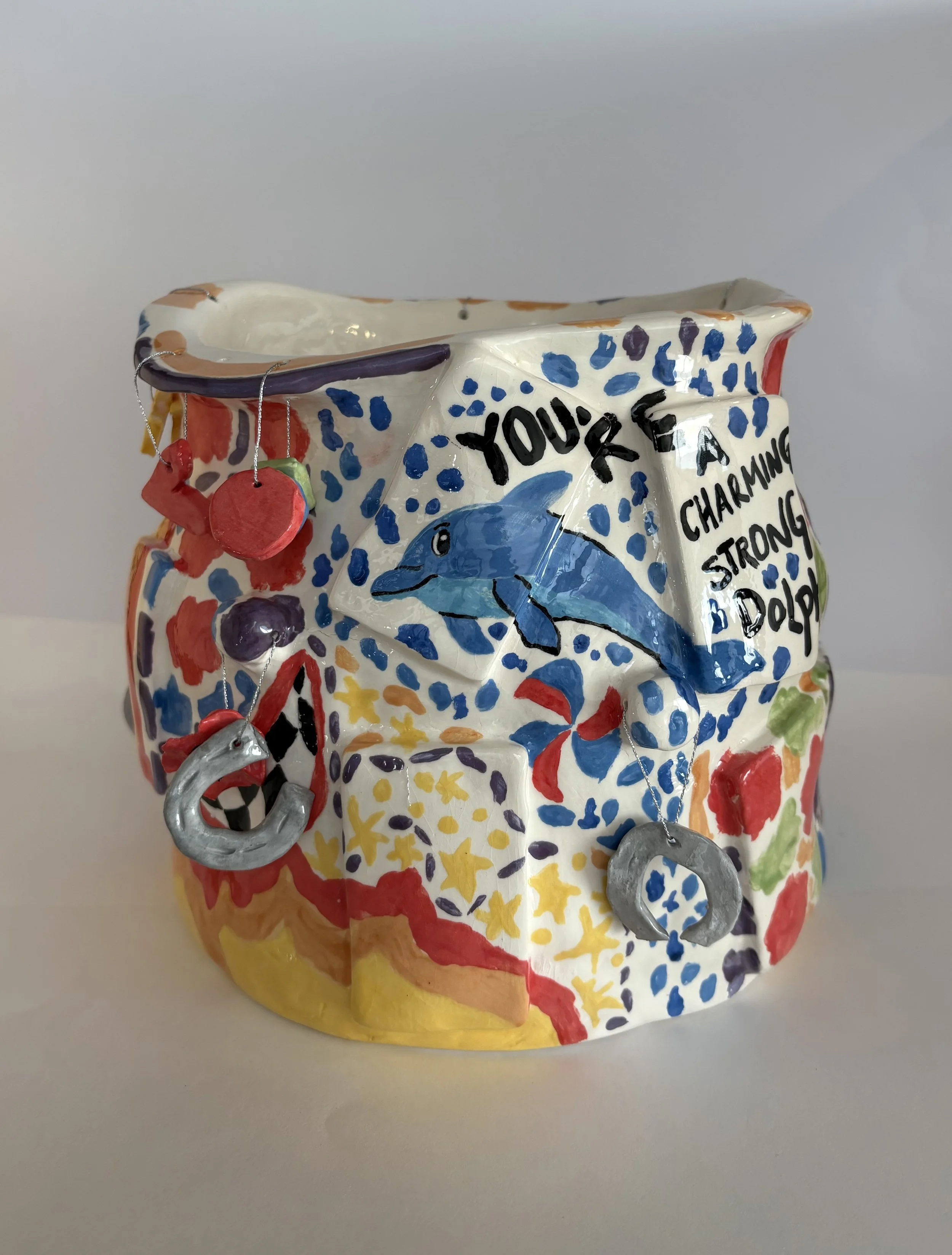 Colourful, eye-catching, earthenware ceramic sculpture, featuring a dolphin, horse shoe charms, and text.