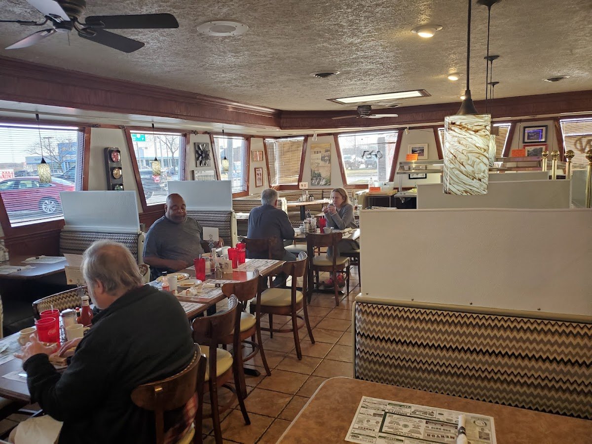 Interior of a casual diner or restaurant with customers sitting at tables, eating, and engaging in conversation. The space has large windows with blinds, various wall decorations, and hanging pendant lights.