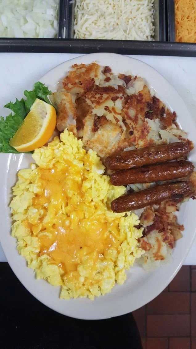 A breakfast plate with scrambled eggs topped with shredded cheese, fried hashbrowns, three cooked breakfast sausage links, a lemon wedge, and a piece of lettuce.