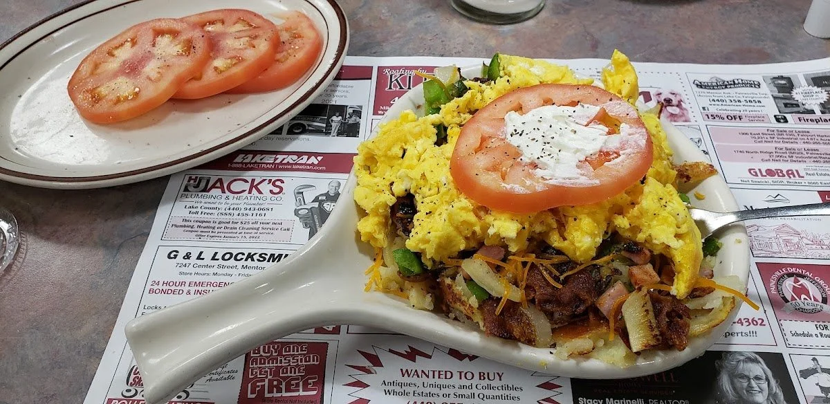 A plate of sliced tomatoes and an elaborate breakfast casserole with eggs, vegetables, and meat, topped with a tomato slice and sour cream, placed on a newspaper-advertising sheet of paper.