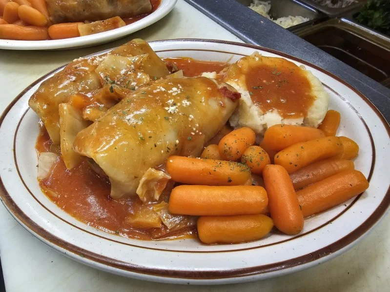 Plate with baked enchiladas topped with cheese and sauce, a serving of mashed potatoes with gravy, and cooked baby carrots.