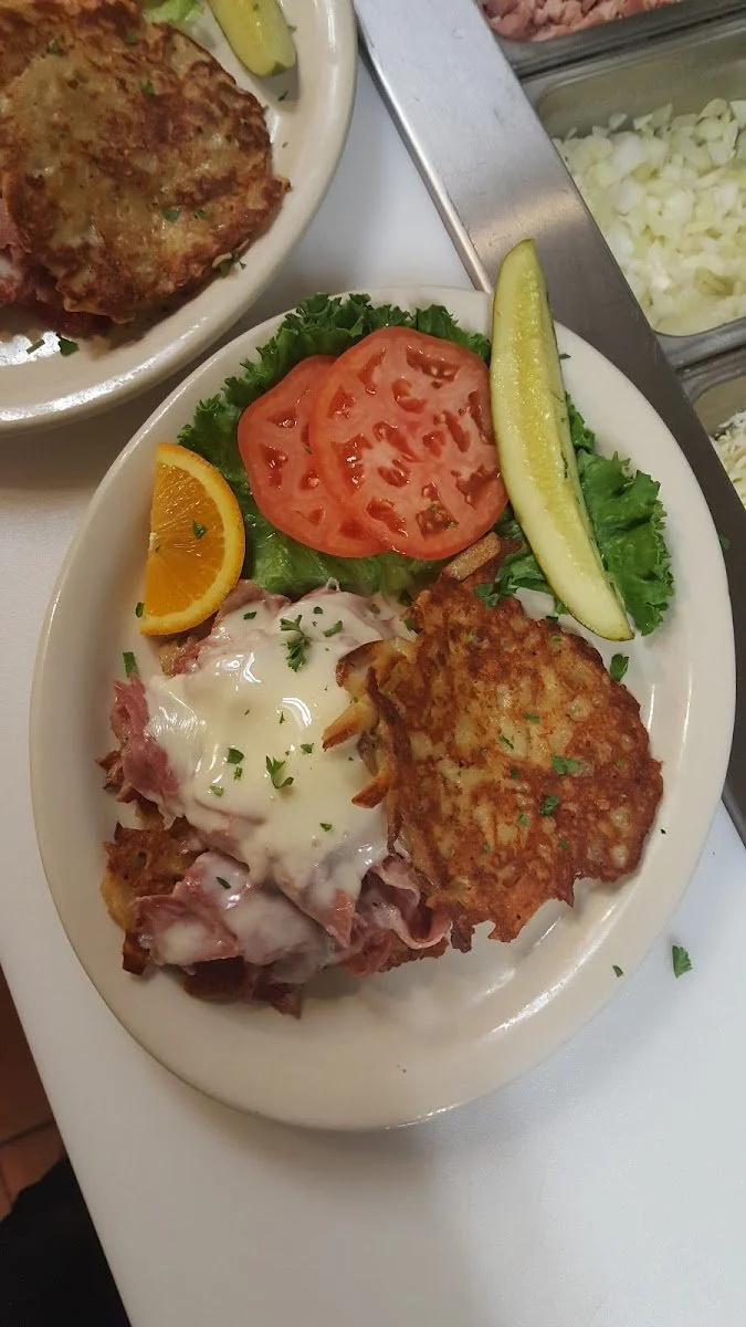 A plate of food with sliced tomatoes, lettuce, a pickle spear, an orange wedge, a serving of meat with gravy and onions, and a crispy potato pancake.