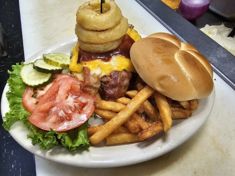 Plate of a bacon cheeseburger with onion rings, lettuce, tomato, pickles, and French fries.