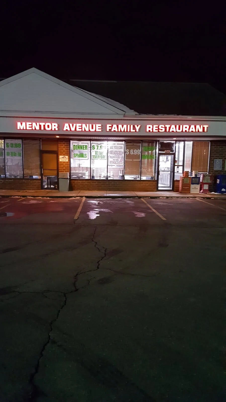 Night view of a restaurant called Mentor Avenue Family Restaurant with illuminated sign, large windows displaying advertisements, and a parking lot with cracked pavement in the foreground.