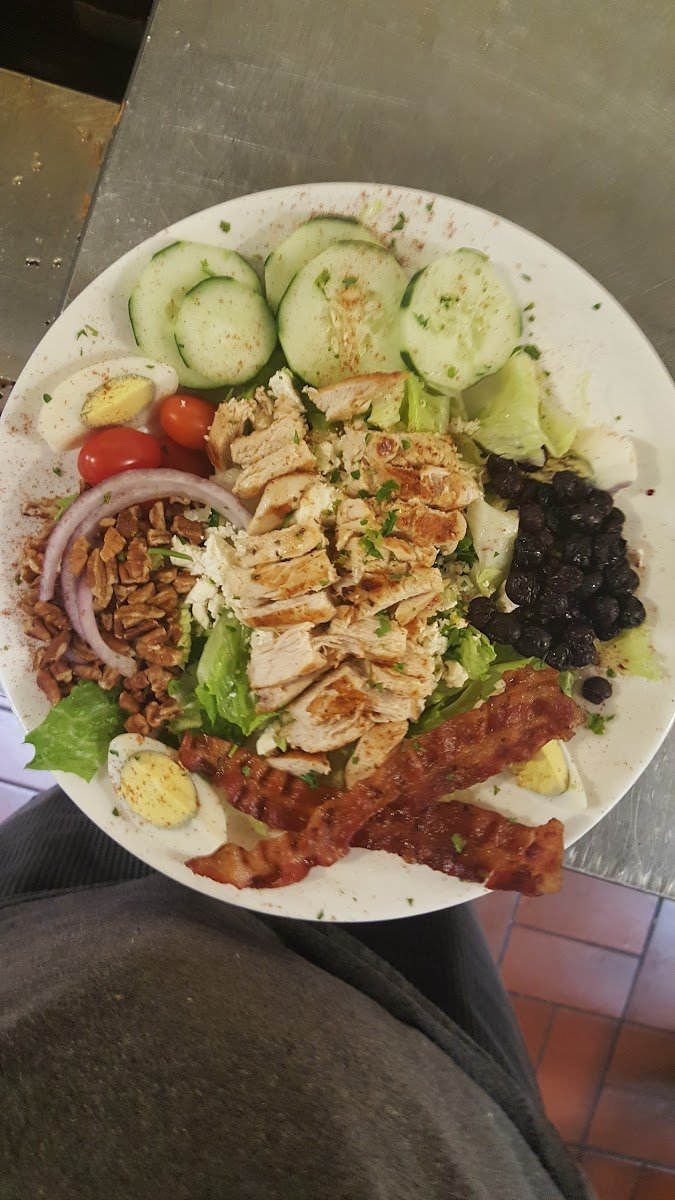 A plate with sliced cucumbers, cherry tomatoes, sliced red onions, chopped nuts, sliced grilled chicken, black beans, bacon, potato salad, and halved boiled eggs, on a metal surface.