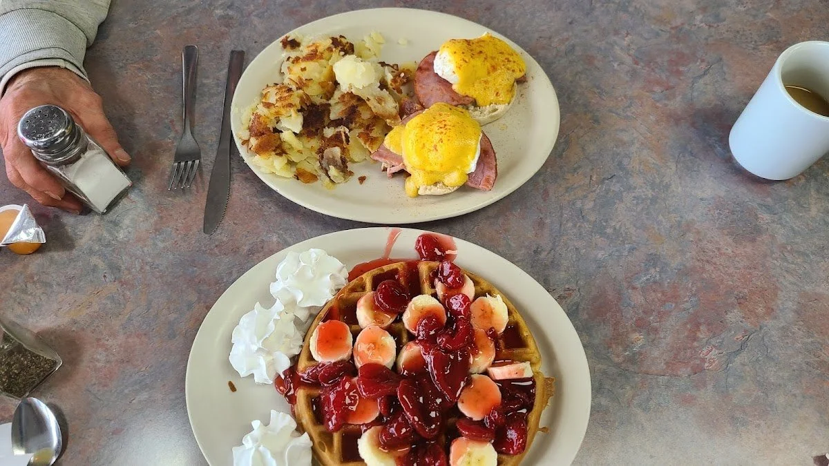 Plate of waffles topped with banana slices, cherry pie filling, and whipped cream, with a side of more whipped cream. A separate plate contains eggs Benedict with ham and poached eggs, served with home fries. A cup of coffee and salt and pepper shake