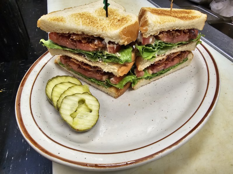 Half of a club sandwich with layers of tomato, lettuce, turkey, and cheese, cut into quarters, served with pickle slices on a white plate with a brown rim.