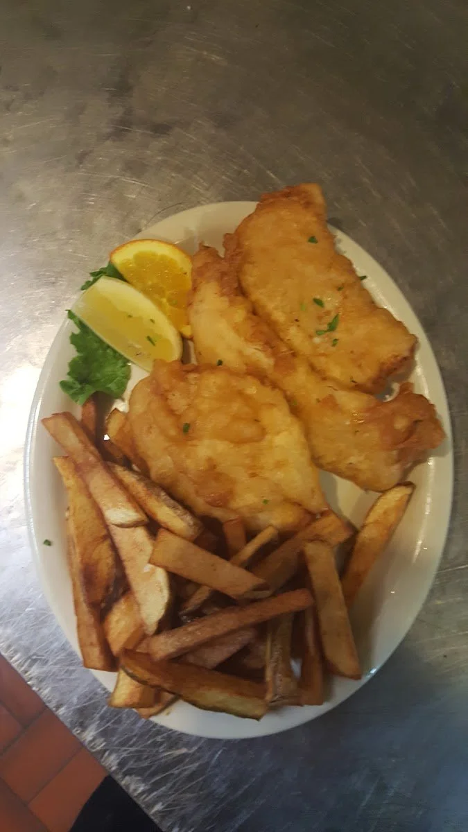 Plate of fried fish, French fries, lemon wedge, orange wedge, and green lettuce leaf on a white oval plate.