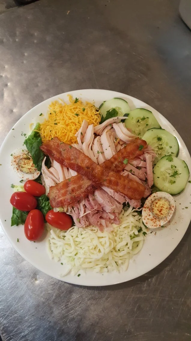 A plate of assorted salad ingredients including shredded cheddar cheese, sliced cucumber, cherry tomatoes, shredded mozzarella cheese, sliced turkey, bacon strips, shredded lettuce, and a halved boiled egg with paprika.