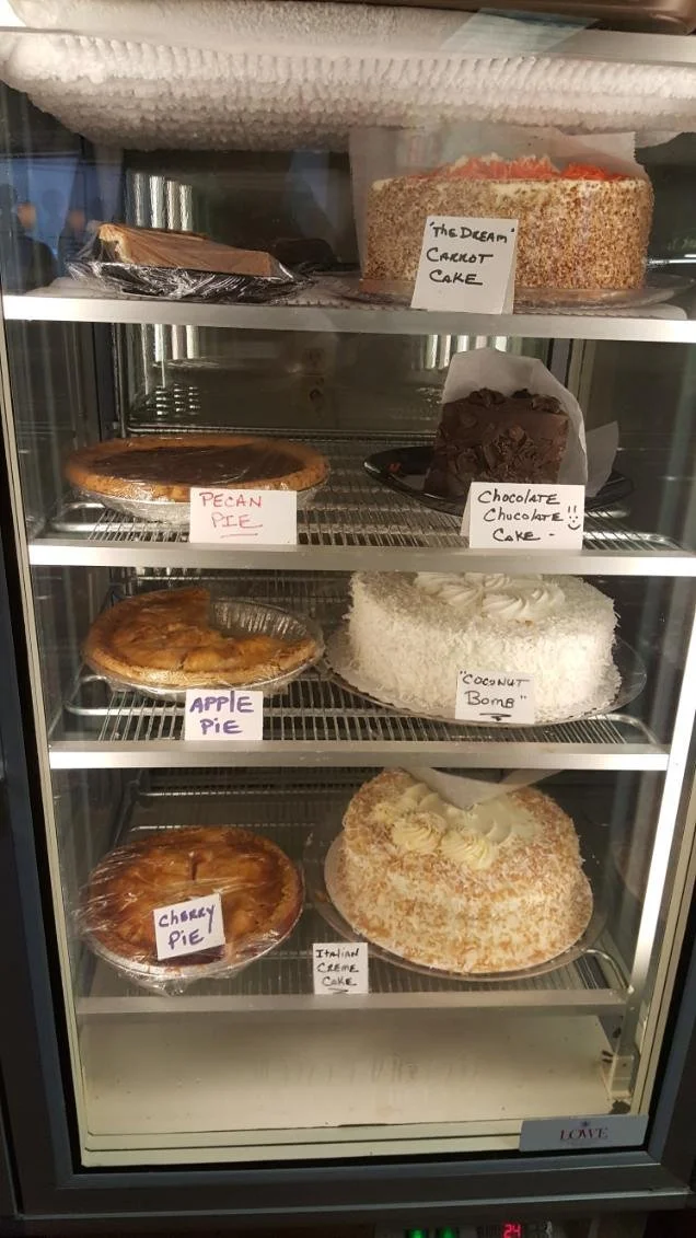 A glass display case filled with various cakes, including a carrot cake labeled 'The Dream Carrot Cake' at the top, a chocolate cake labeled 'Chocolate Chocolate!! Cake', a pecan pie labeled 'Pecan Pie', an apple pie labeled 'Apple Pie', a cherry pie