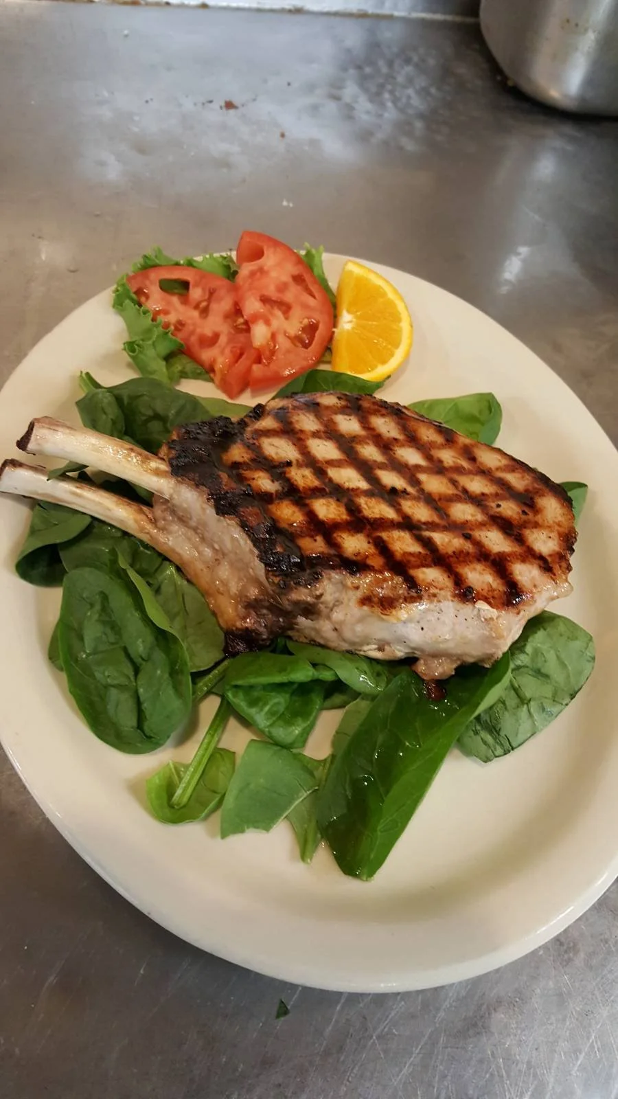 Grilled pork chop on a bed of spinach, with sliced tomato, lemon wedge, and lettuce on a white plate.