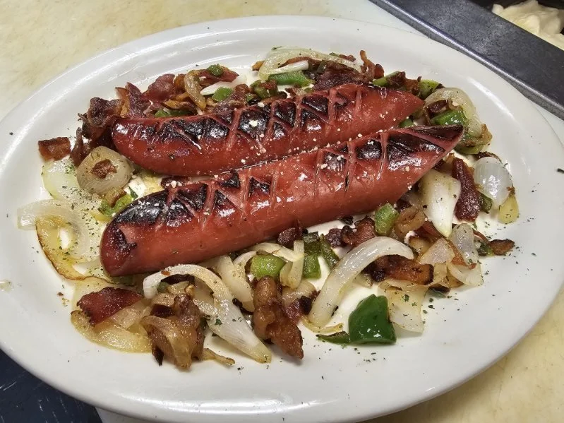 Grilled sausages on a plate with sautéed onions, green peppers, and bacon.