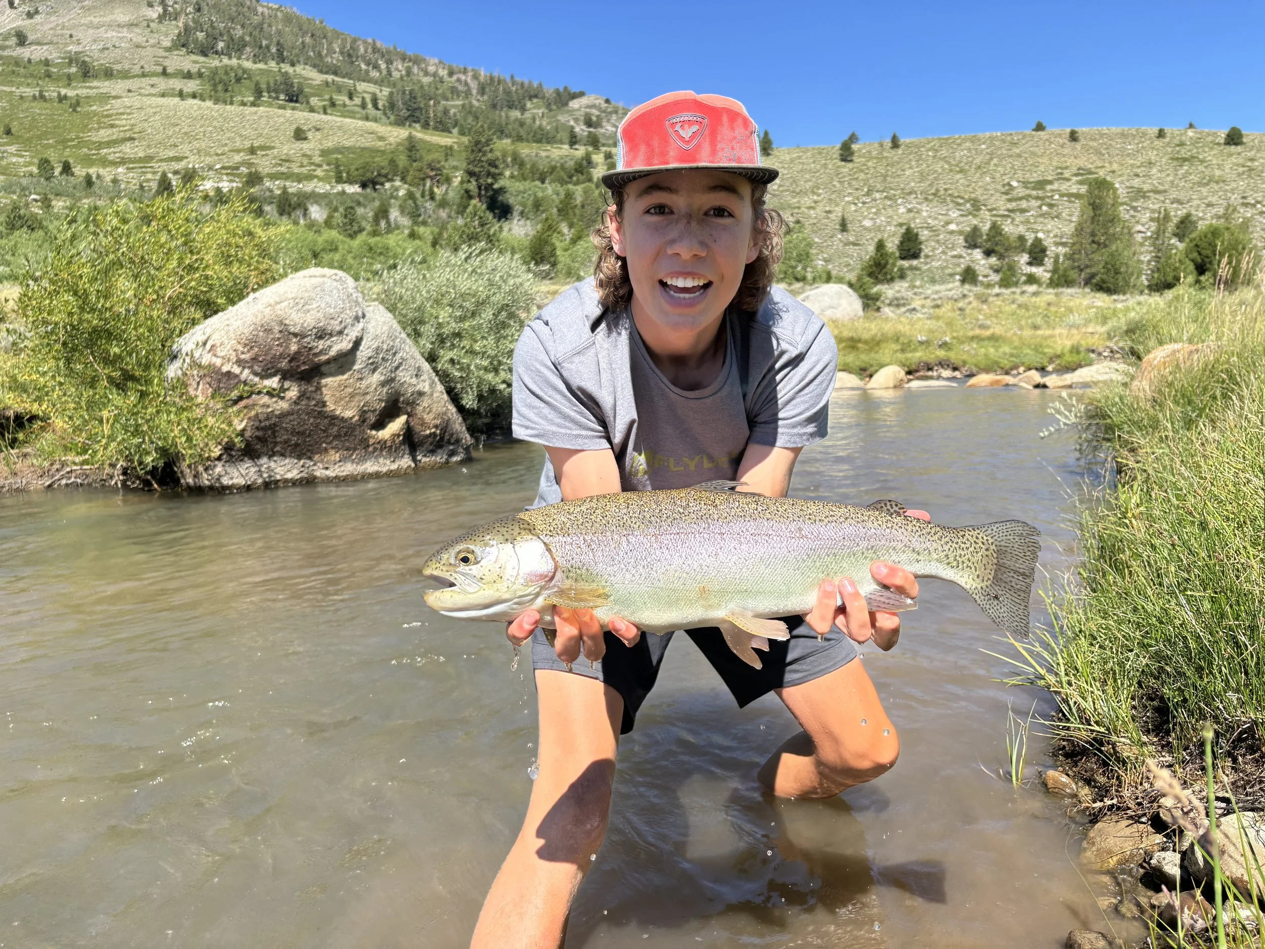 A person kneeling in a river, holding a large fish they've caught. The person is wearing a red hat and gray shirt, and the background features green hills and a clear blue sky.