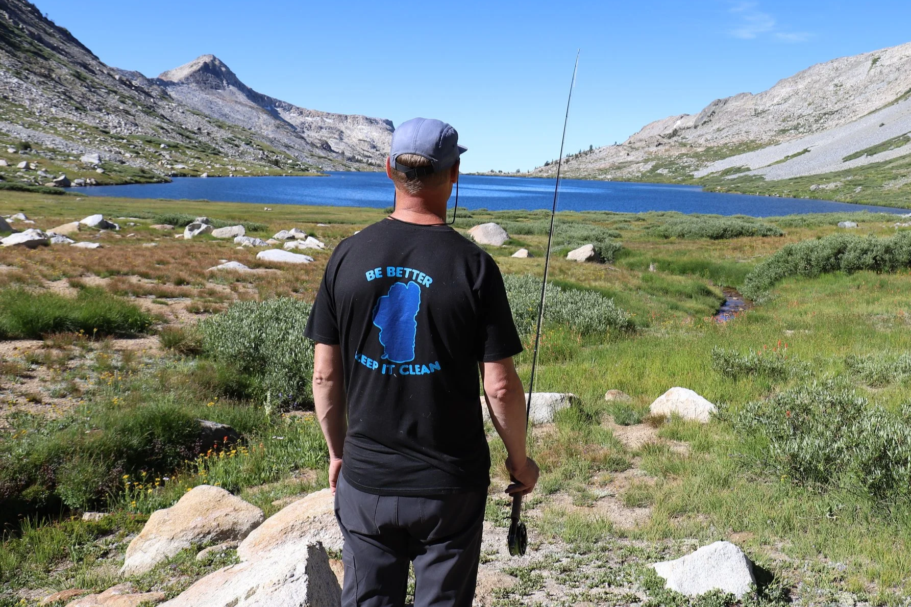 A person standing outdoors near a lake and mountains, holding a fishing rod, wearing a black t-shirt with blue text and logo, a gray cap, and gray pants.