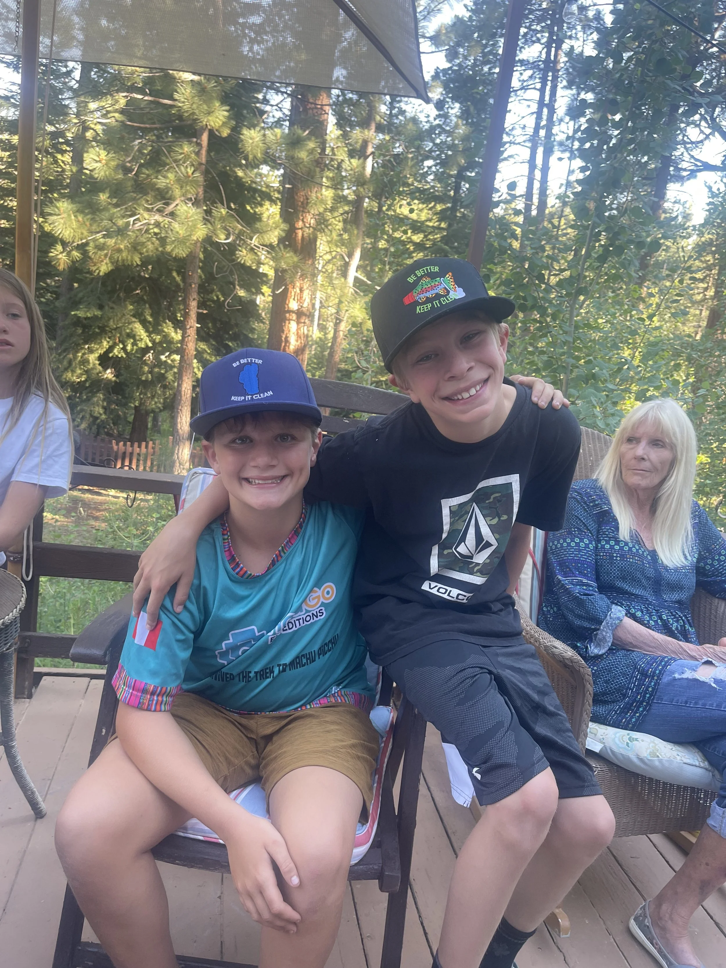A group of young boys and a woman sitting outdoors in a forested area with tall trees, some sitting on chairs, in casual clothing, enjoying a sunny day.