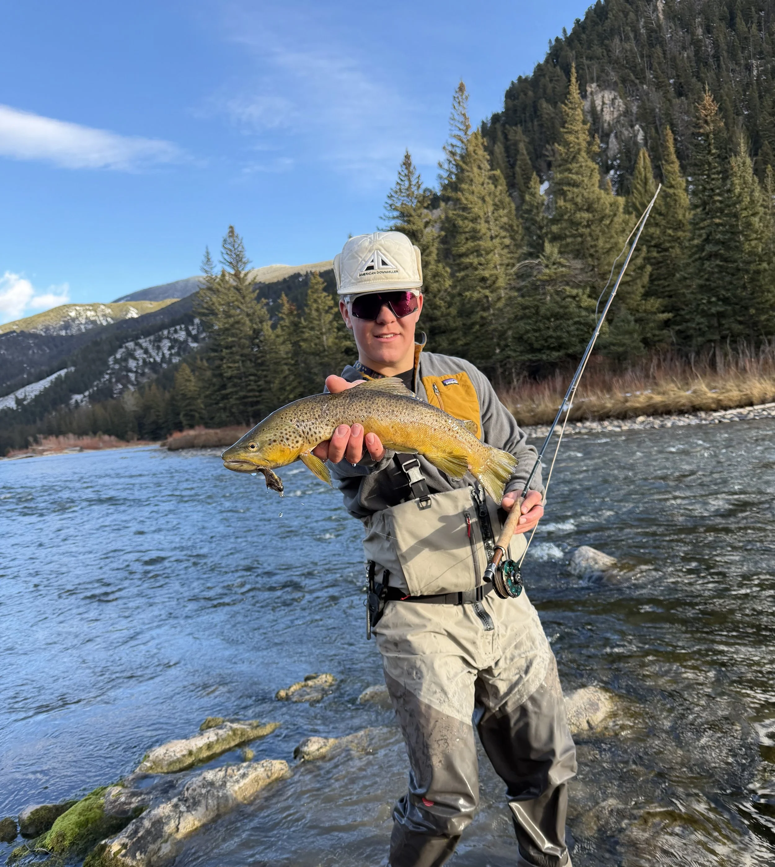 Person holding a large fish while standing in a river, surrounded by snow-capped mountains and evergreen trees.
