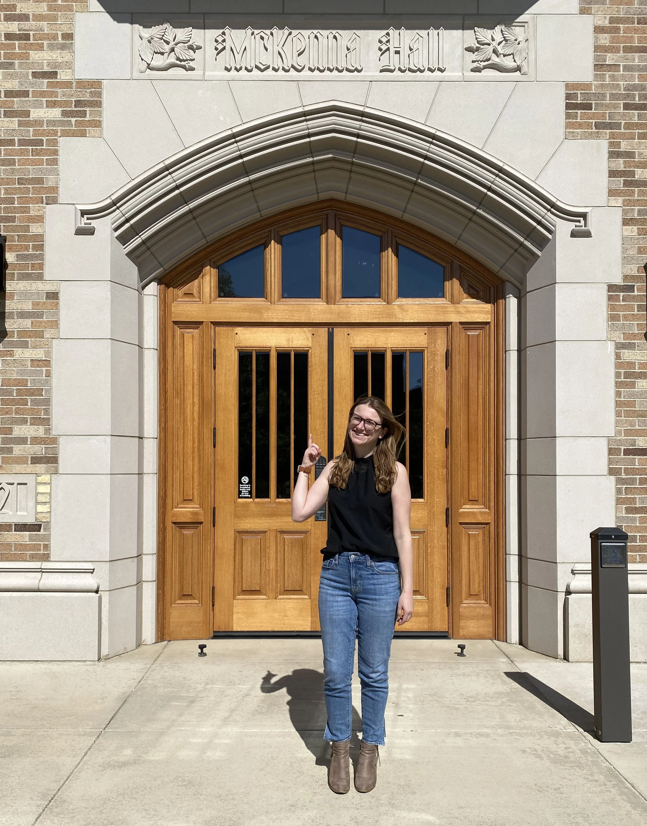 A woman with glasses,  standing in front of a large wooden door with an arched stone entryway labeled 'McKenna Hall'.