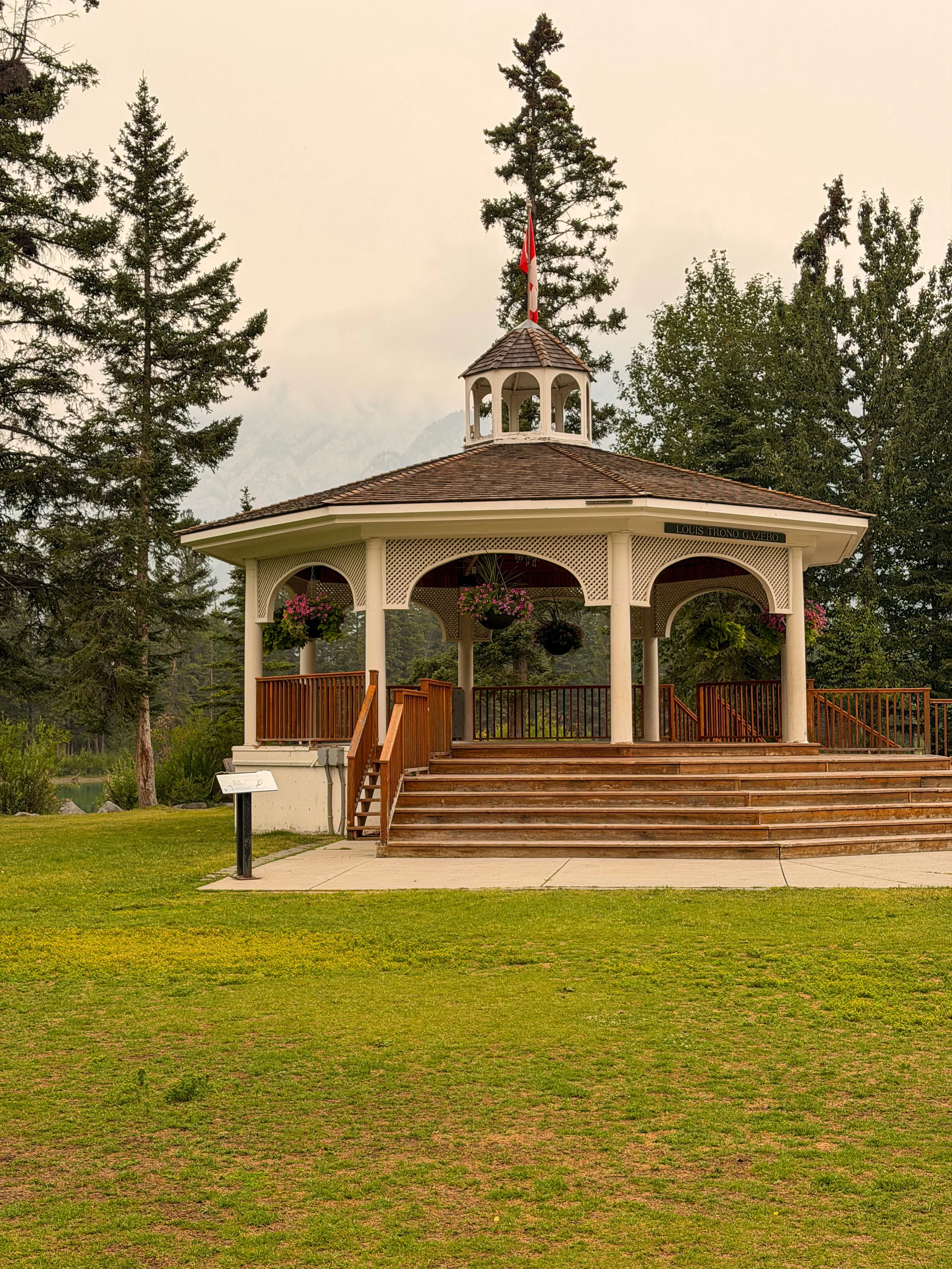 A gazebo with a wooden staircase and railings, hanging flower baskets, and a flagpole with a Canadian flag, surrounded by tall trees and mountains in the background.