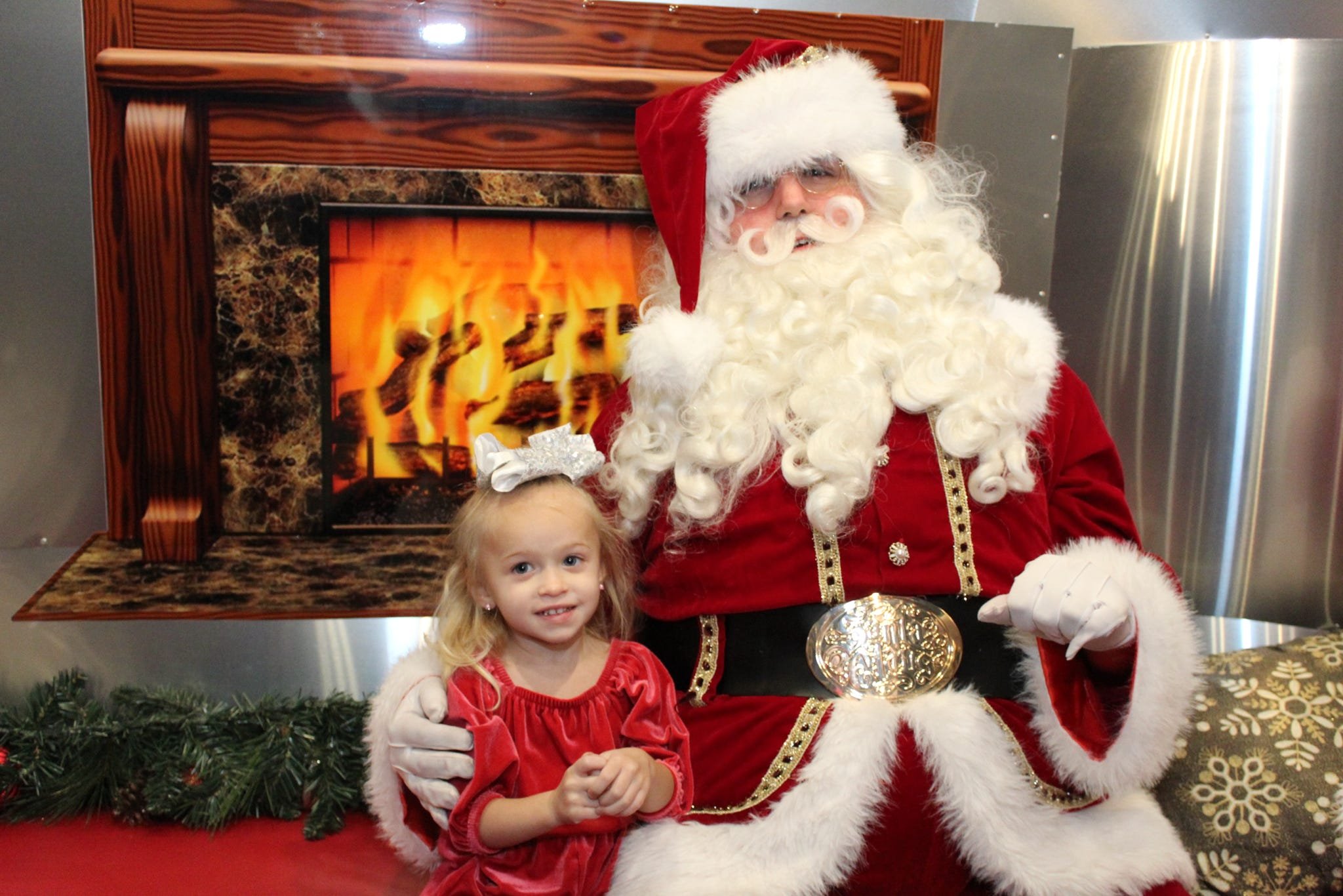 A young girl in a red dress sitting with Santa Claus, who is dressed in a traditional red suit with white fur trim, in front of a faux fireplace with a fire burning. The girl has blonde hair and is looking at the camera. Santa has a long white beard, glasses, and is holding a gold ornament in his gloved hand. There is festive greenery and wrapped presents nearby.