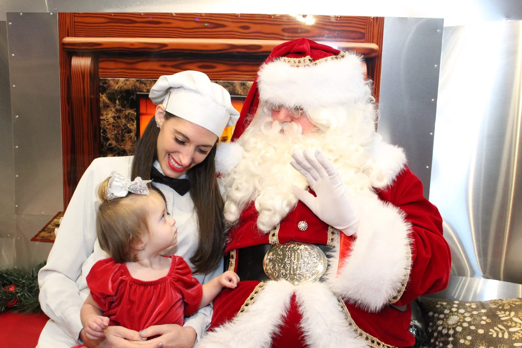 A woman dressed as Santa's helper sitting with two children, smiling as Santa Claus, in his red suit and hat, interacts with them in a festive setting.