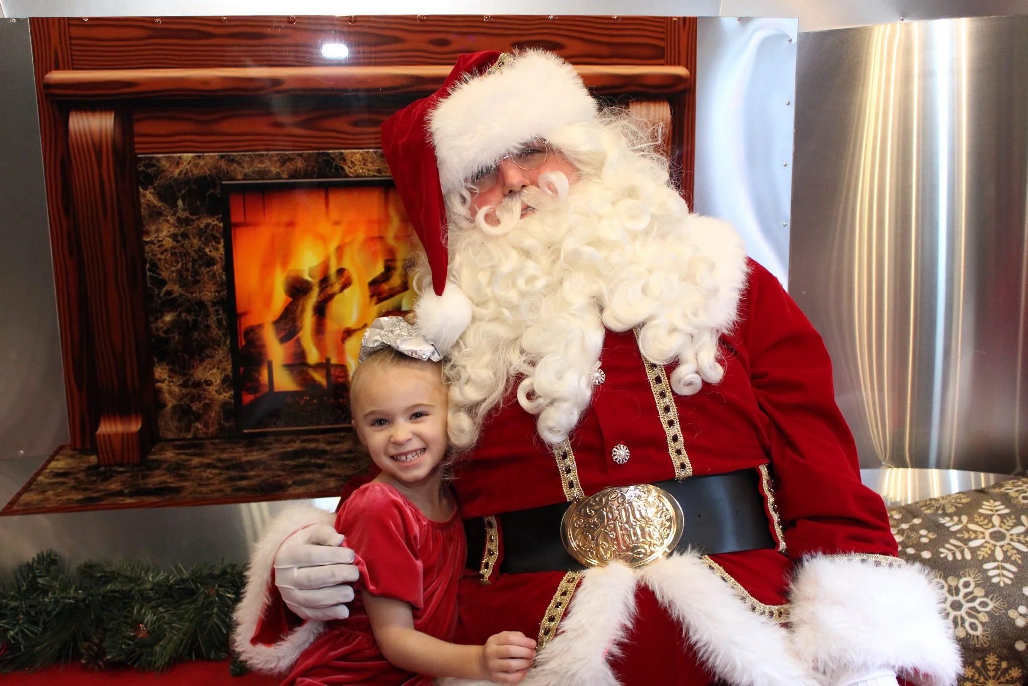 A young girl in a red dress with white fur trim standing next to Santa Claus, who is wearing a red suit with white fur trim, a red Santa hat, and a large gold belt. They are sitting in front of a fireplace with a glowing fire and a wooden mantel. Santa has a white curly beard and glasses. The girl is smiling, showing her teeth, and has a bow in her hair. The setting is festive with holiday decorations.