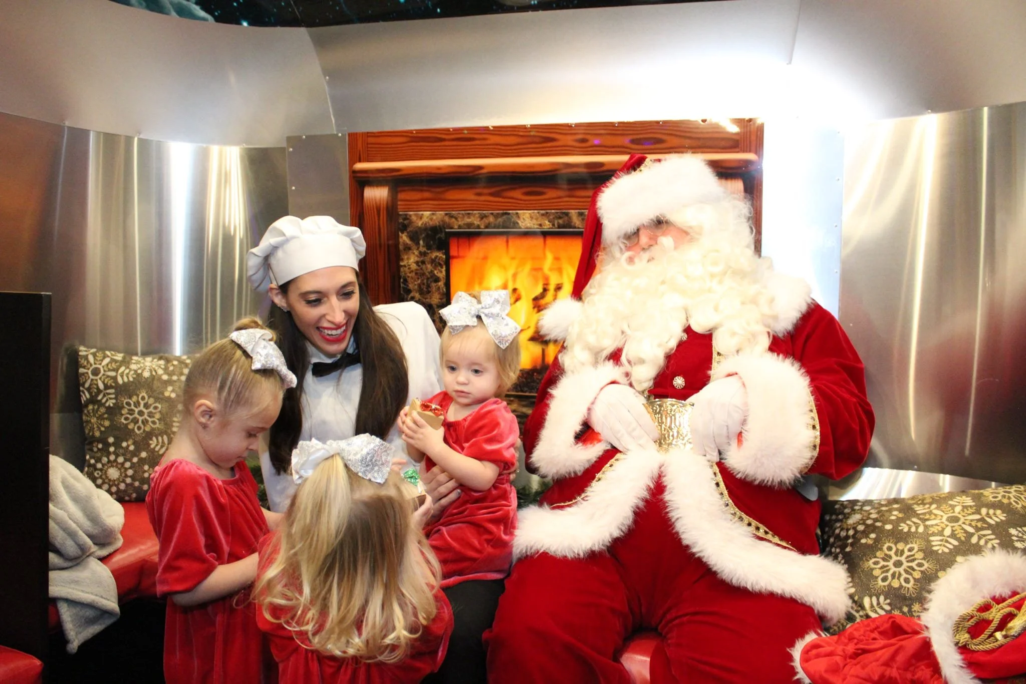 Santa Claus dressed in his traditional red suit with white fur trim, sitting with children dressed in Christmas-themed red dresses, in front of a fireplace, in a cozy indoor setting.