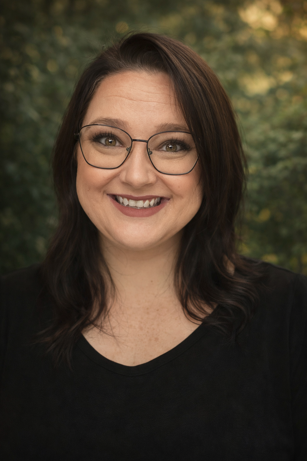 A smiling woman with shoulder-length dark hair and glasses, wearing a black top, outdoors with blurred green foliage in the background.