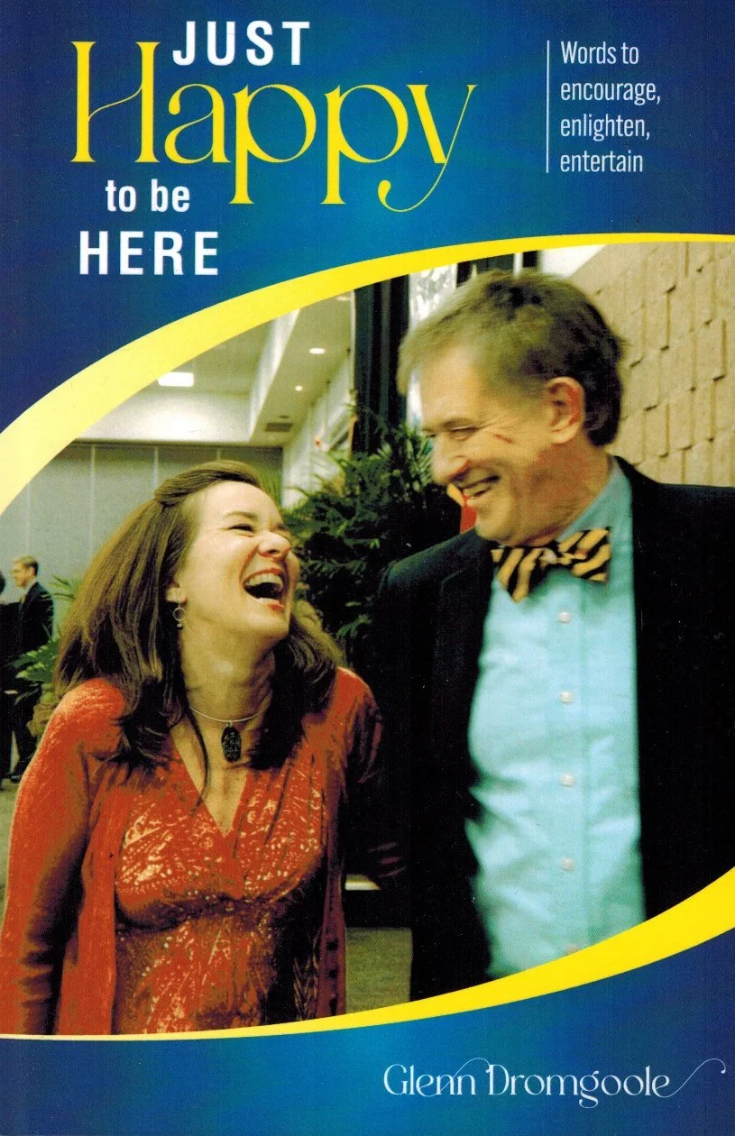 Cover of the book titled "Just Happy to Be Here" by Glenn Dromgoole, featuring a photograph of a woman and a man laughing together, with the woman wearing a red patterned top and the man wearing a light blue shirt with a striped bow tie, indoors with plants and other people in the background.