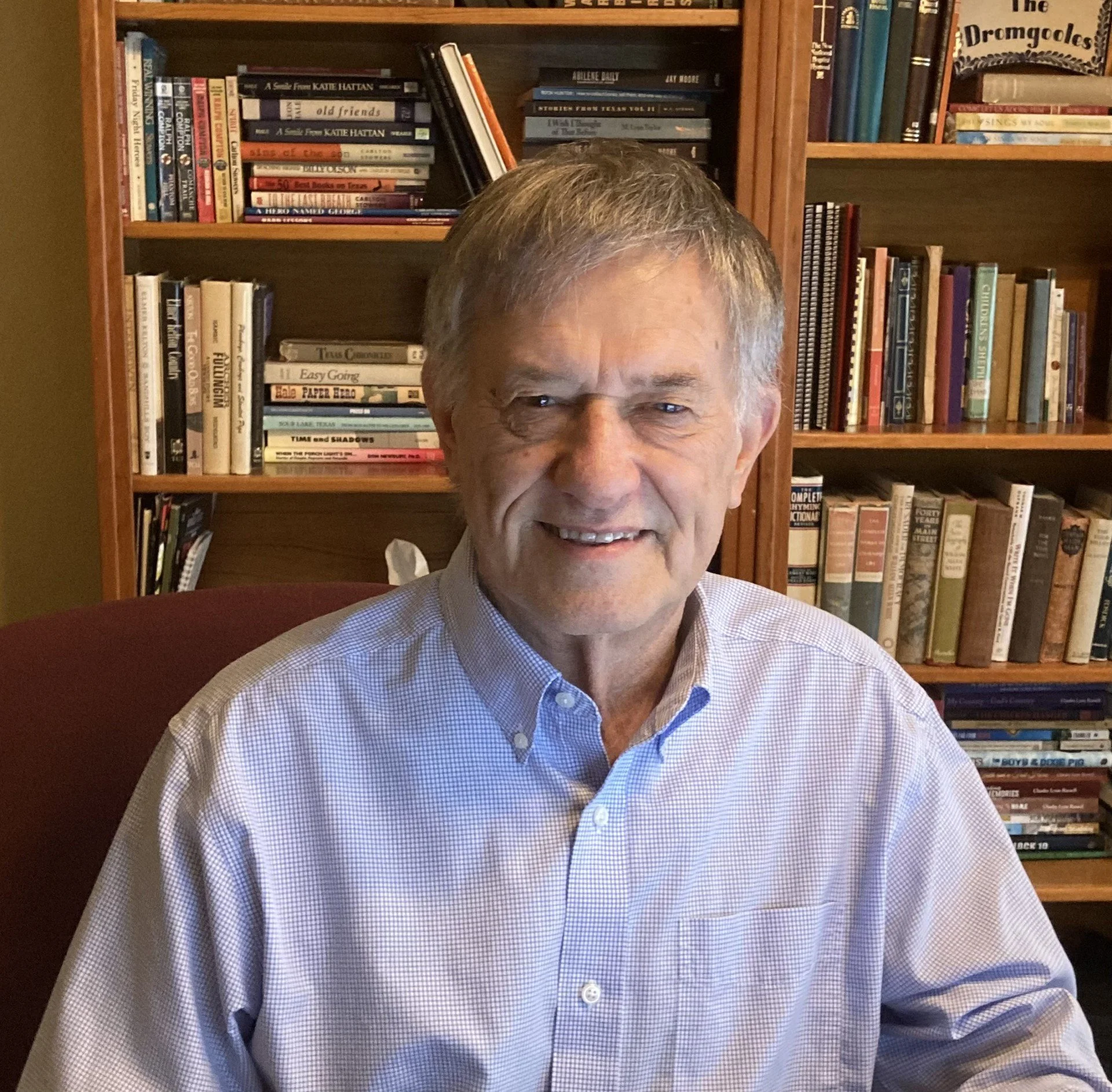 Close-up of an older man with gray hair and a checkered dress shirt, smiling, seated in front of a wooden bookshelf filled with various books.