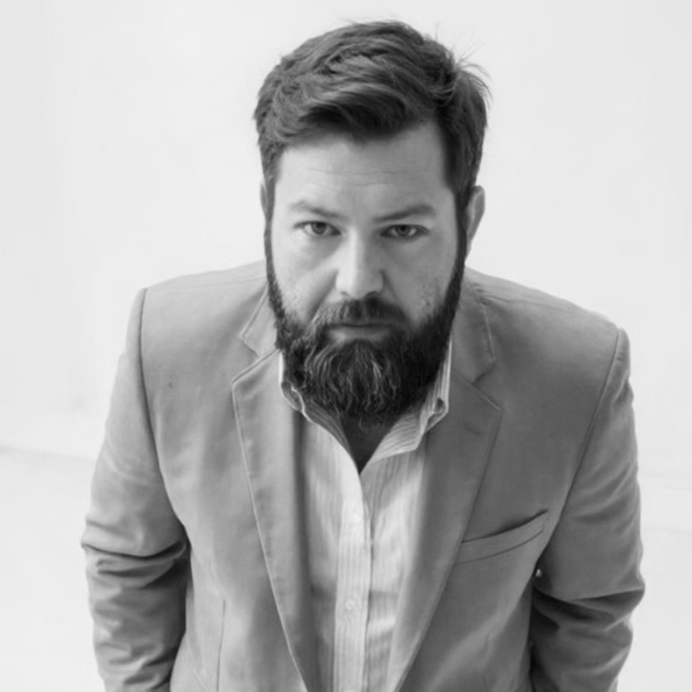 Black and white portrait of a man with a beard wearing a suit jacket and a collared shirt, looking directly at the camera.