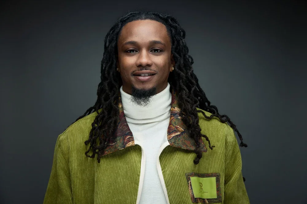 Portrait of a man with long dreadlocks wearing a green jacket and white turtleneck sweater against a dark background.